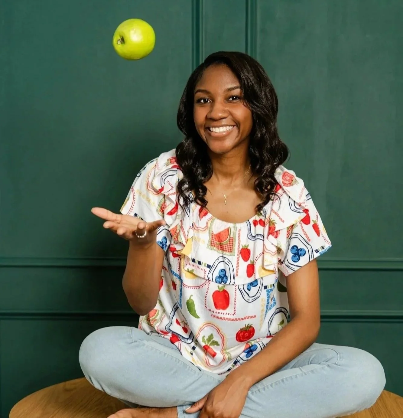 Georgia-based Registered Dietitian Destiney Mallett sitting cross-legged on a wooden surface, smiling and tossing a green apple into the air, in front of a dark green wall.