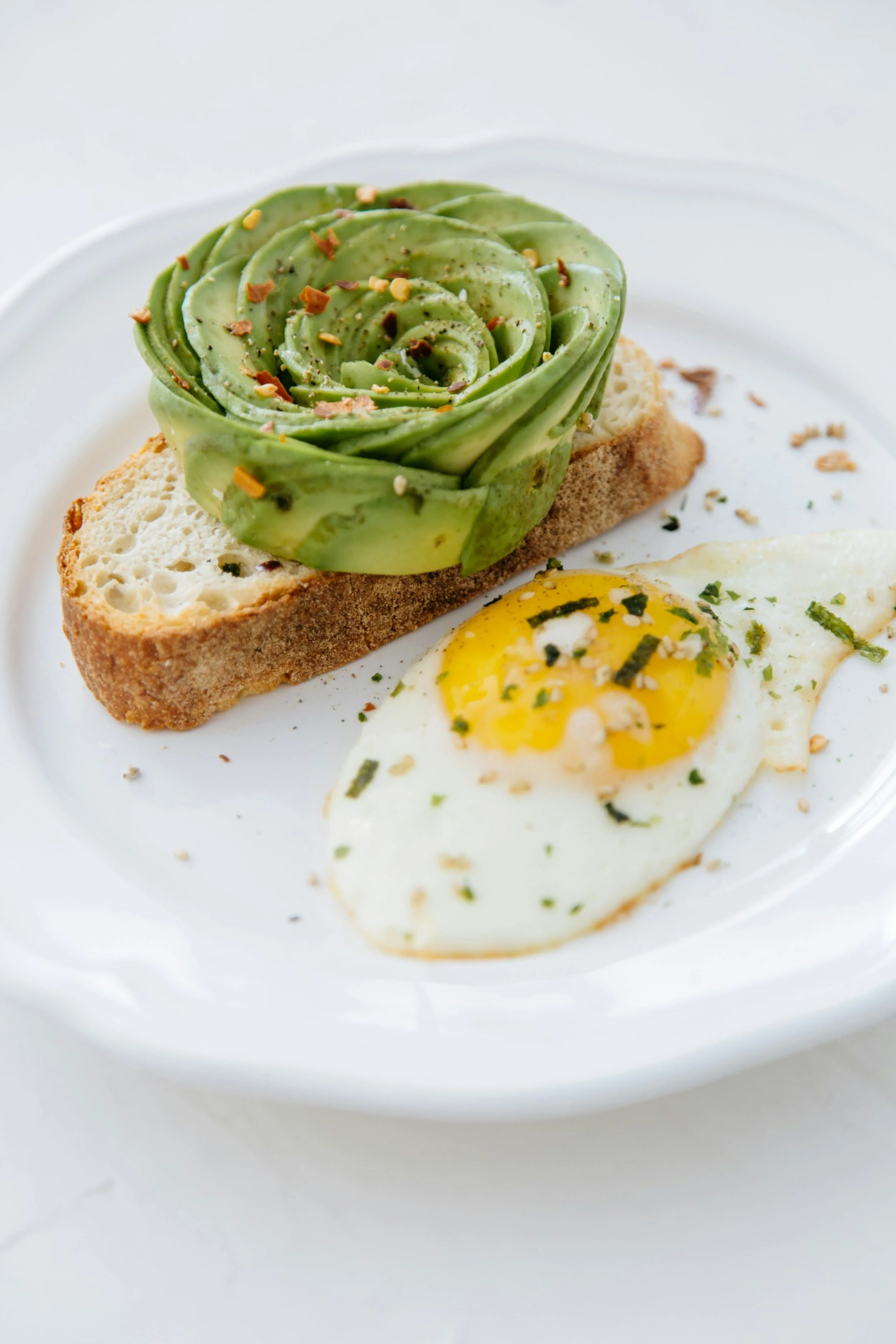 Open-faced sandwich with avocado rose on bread, served with a fried egg garnished with herbs on a white plate.