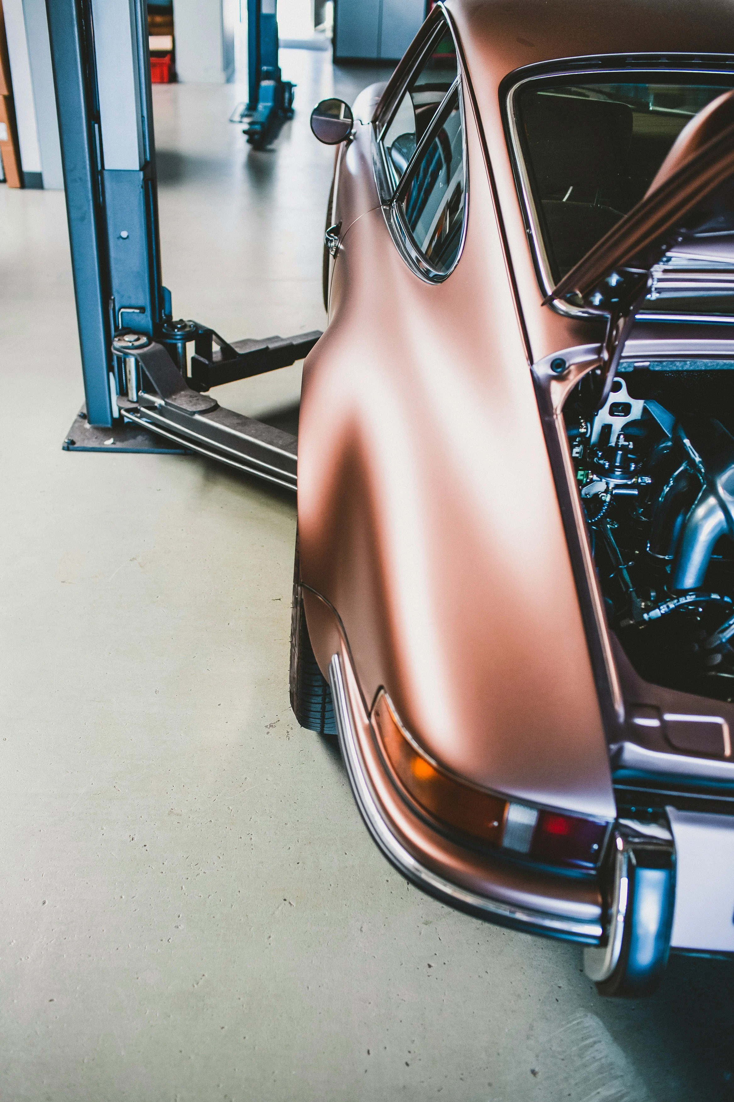 A vintage rose gold-colored car is in a garage, with part of it lifted by a hydraulic lift. The garage has a concrete floor and various tools and storage units are visible in the background.