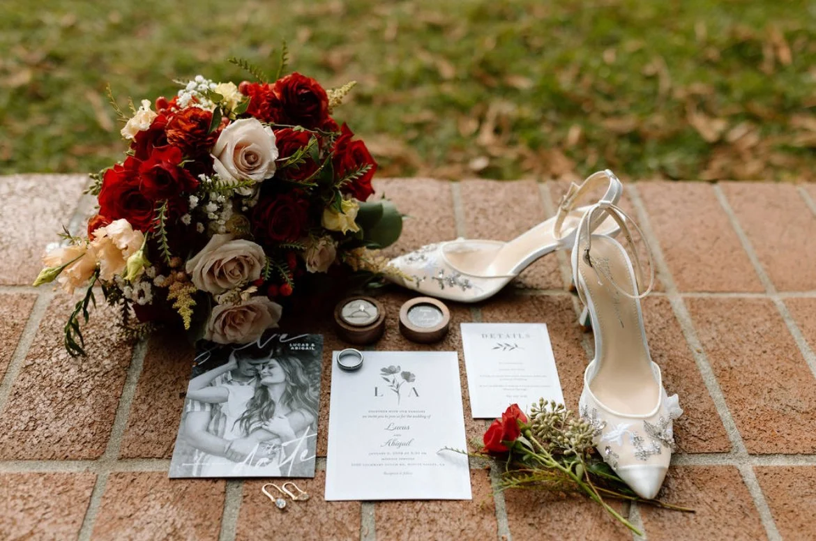 Wedding bouquet with red and white roses, a pair of silver wedding rings, a pair of white high heels with decorative silver details, a wedding invitation, and a photo of the bride and groom, arranged on a brick surface.