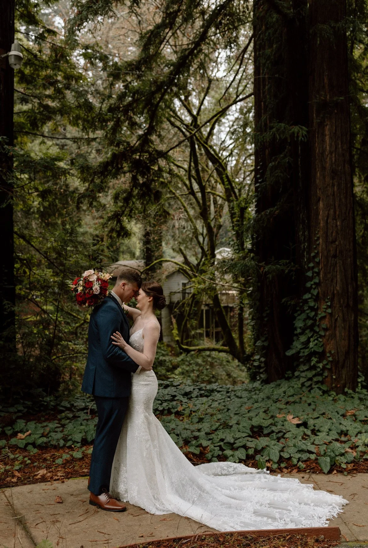 A bride and groom stand close together on a wooded trail surrounded by tall trees and green foliage. The groom in a navy suit holds a bouquet of red and white flowers, while the bride in a white lace gown touches his face. Their foreheads touch as they smile at each other in a romantic moment.