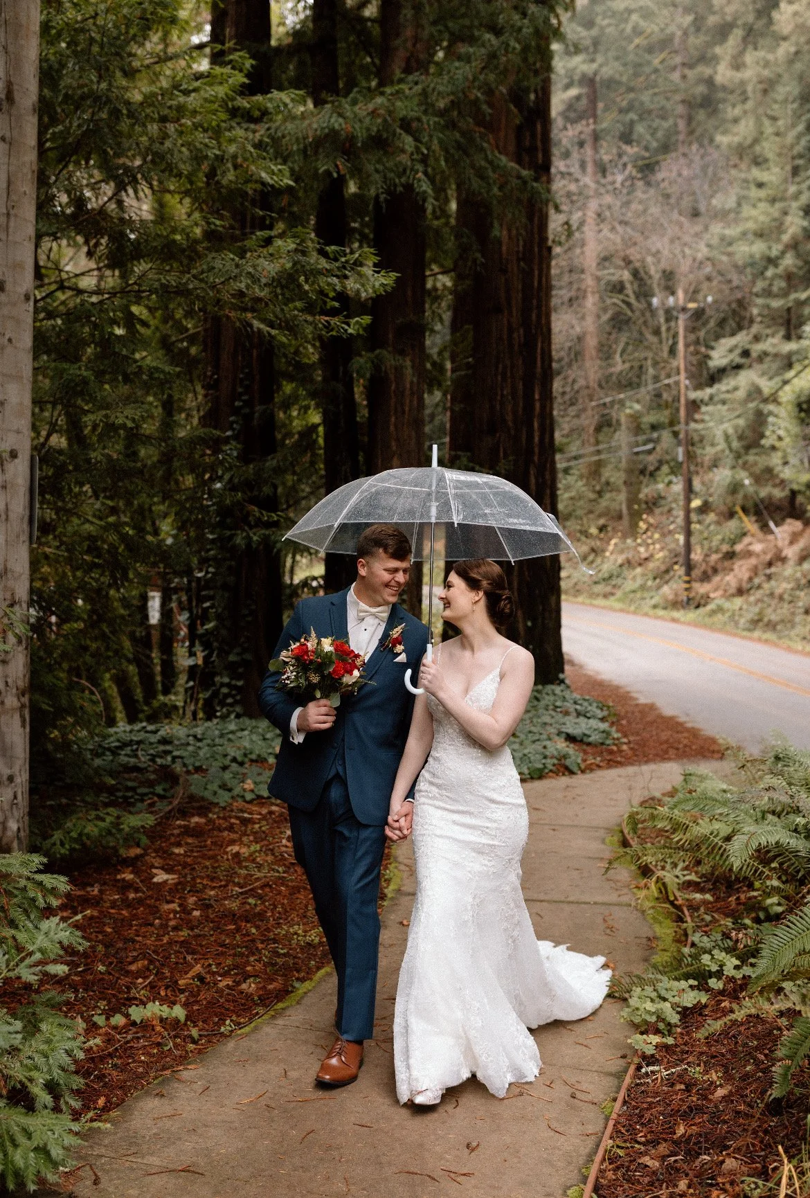 A bride and groom smiling at each other while walking hand in hand on a sidewalk in a wooded area, under an umbrella, with the groom holding a bouquet of flowers.
