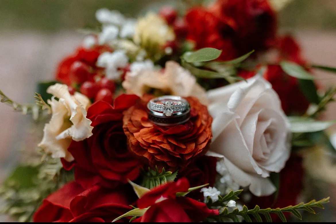 Close-up of a wedding ring and engagement ring placed on a bouquet of red, white, and orange flowers with green leaves.