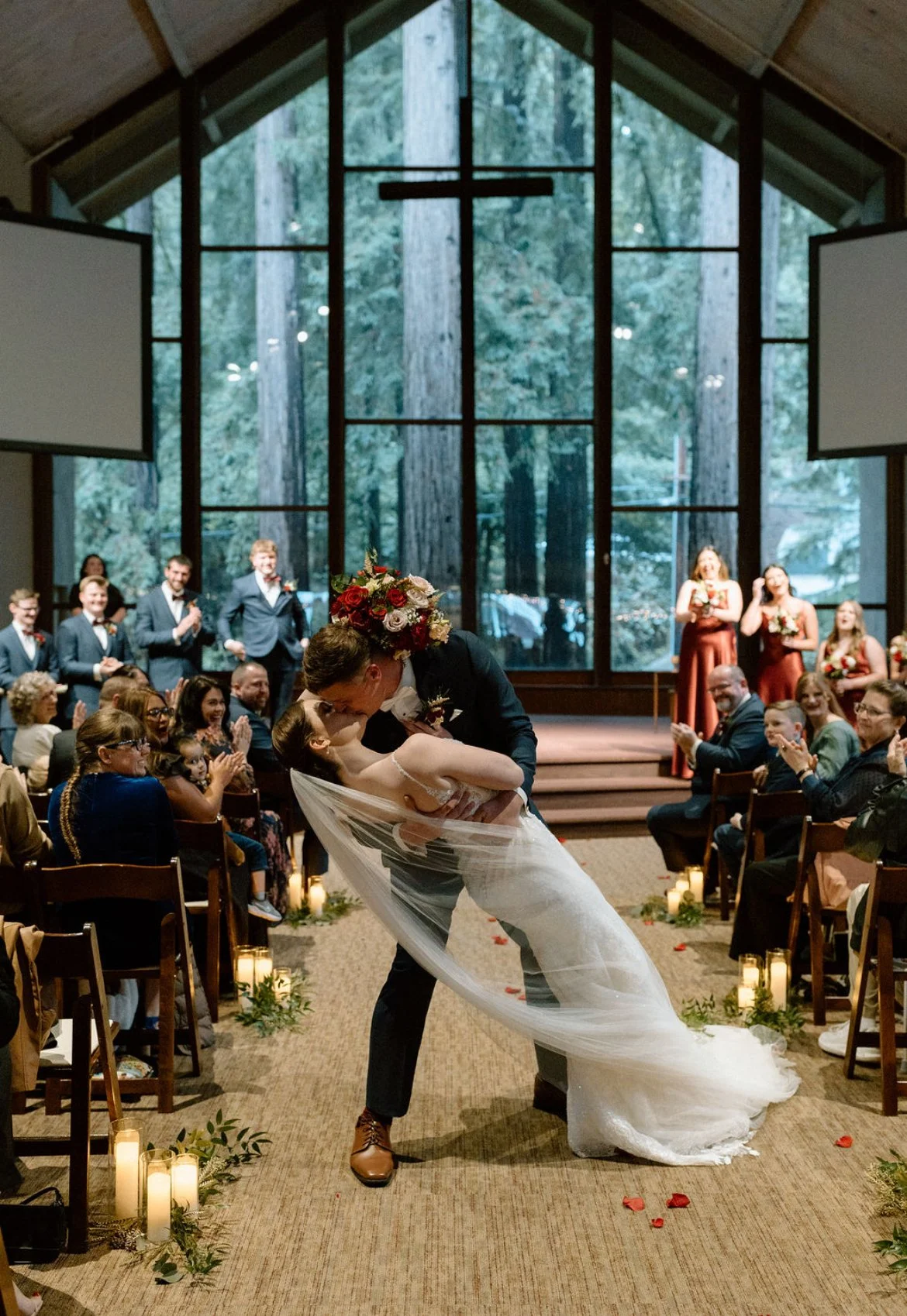 A bride and groom sharing a kiss during their wedding ceremony inside a church, with guests seated on either side and a cross on the large window behind them.