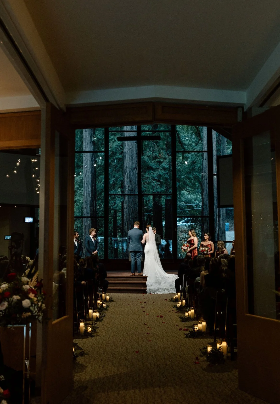 A wedding ceremony taking place inside a church or chapel with guests seated on either side of the aisle, decorated with candles and flowers, and a large window with trees in the background behind the altar.
