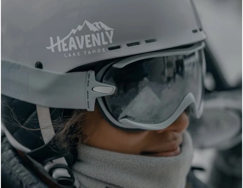 Close-up of a skier wearing a gray helmet with 'Heavenly Lake Tahoe' logo, ski goggles, and a gray neck warmer, in a snowy environment.