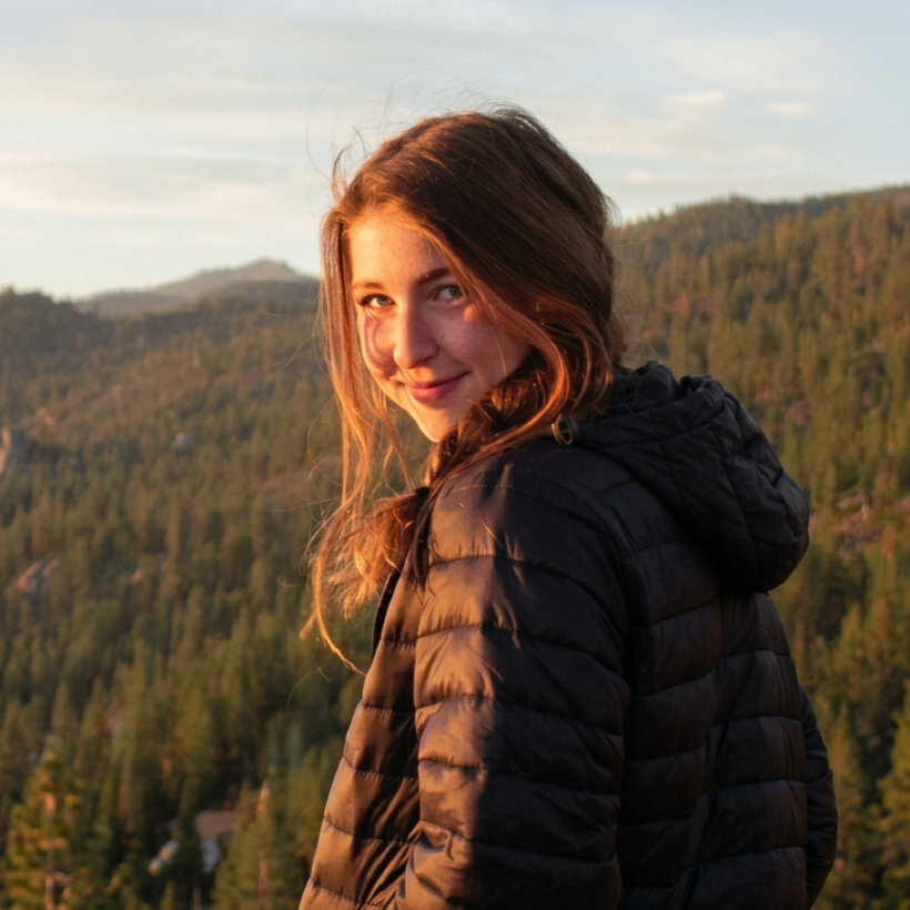 A young woman with long wavy brown hair smiling at the camera, wearing a black puffy jacket, with a forested mountain landscape in the background during sunset.