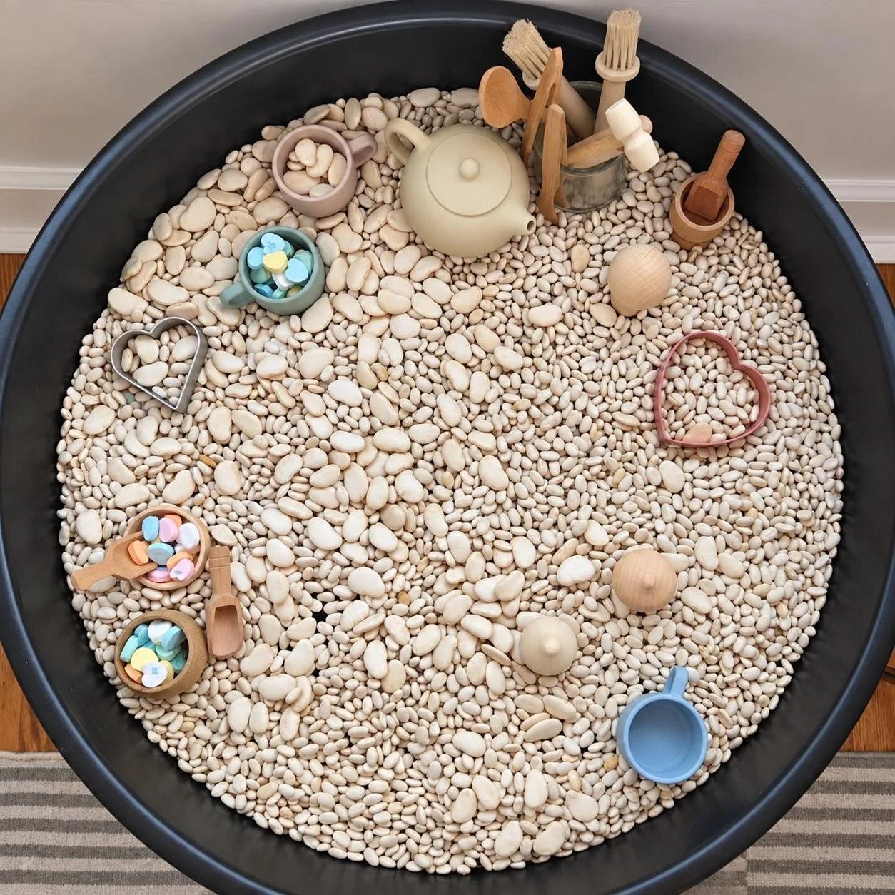 A large sensory bin filled with white pebbles, containing small toys including heart-shaped cookie cutters, wooden scoops, a teapot, and small containers for scooping and pouring at the little learning center in Norwalk, CT.