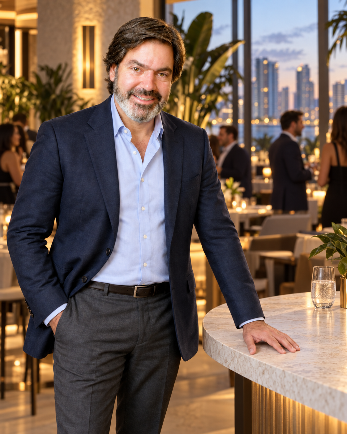 A middle-aged man with brown hair and a beard, wearing a blue blazer and gray pants, standing at a marble-topped bar in a modern, upscale restaurant or lounge. The background shows several people socializing and city skyline views through large windows during evening.