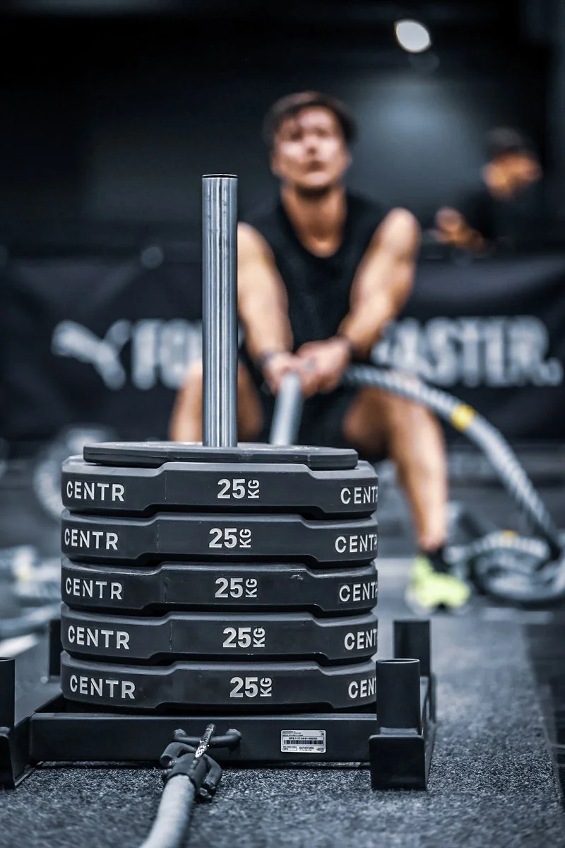 A Hyrox athlete in workout gear is in a gym pulling a battle rope, with a stack of 25 kg weight plates in the foreground.