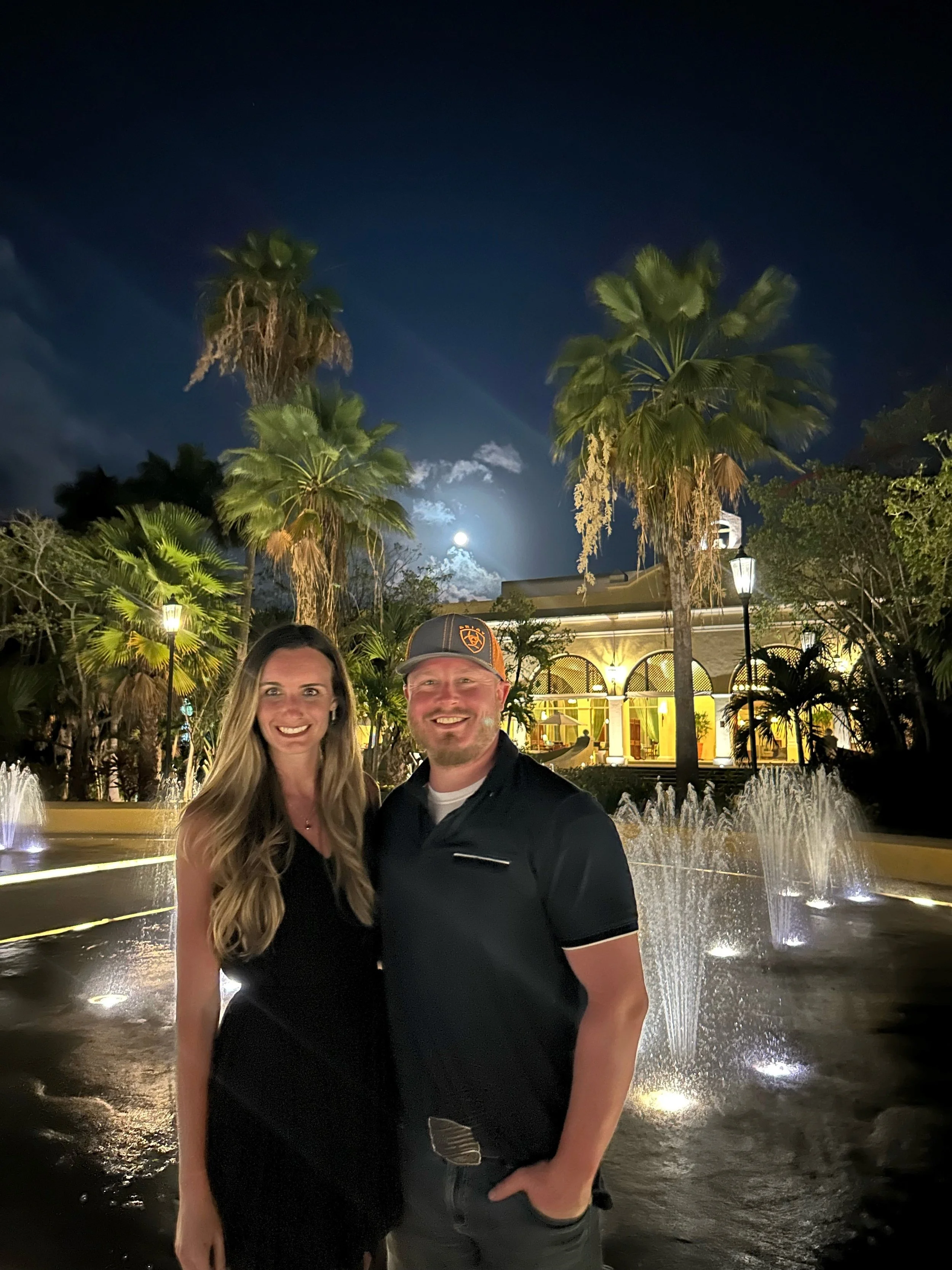 A smiling man and woman standing together in front of a fountain at night, with palm trees and a building illuminated in the background.