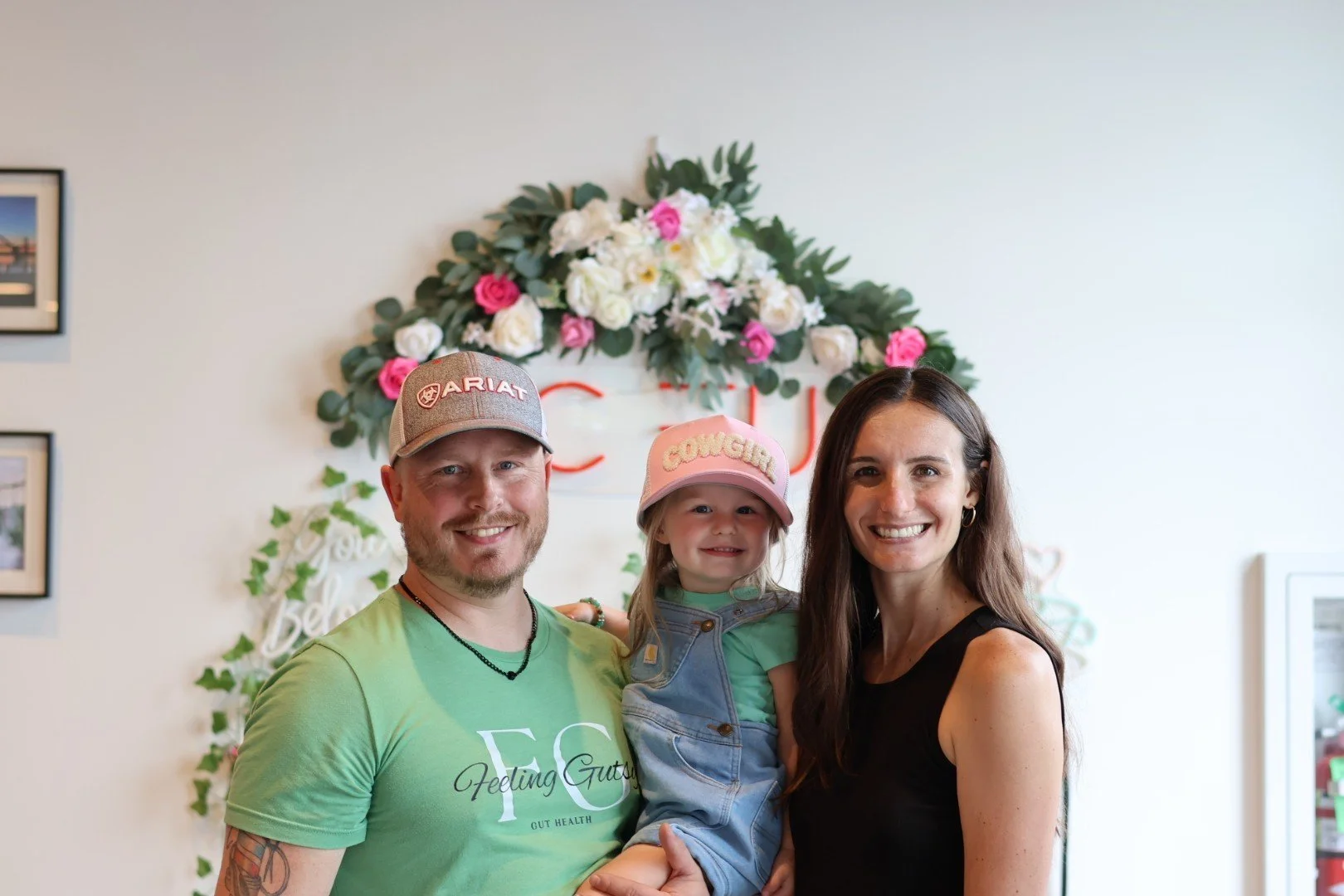 A family of three smiling for a photo indoors, with a floral decoration and a sign that says "Cocu" in the background. The man wears a baseball cap and a green T-shirt, the woman wears a black sleeveless top, and the young girl wears a pink cap and overalls.
