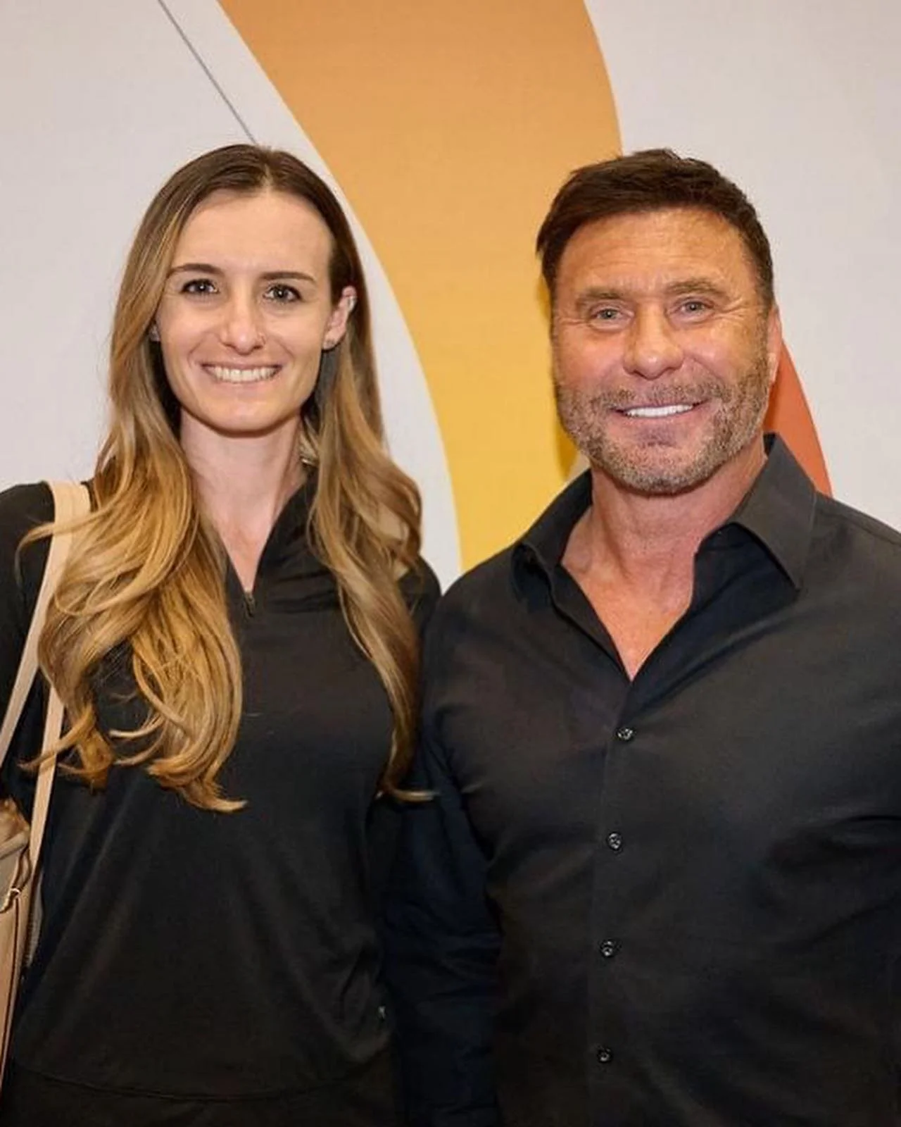A smiling woman with long reddish blonde hair and a man with short dark hair and a beard, both dressed in black, standing together indoors against a colorful abstract background.