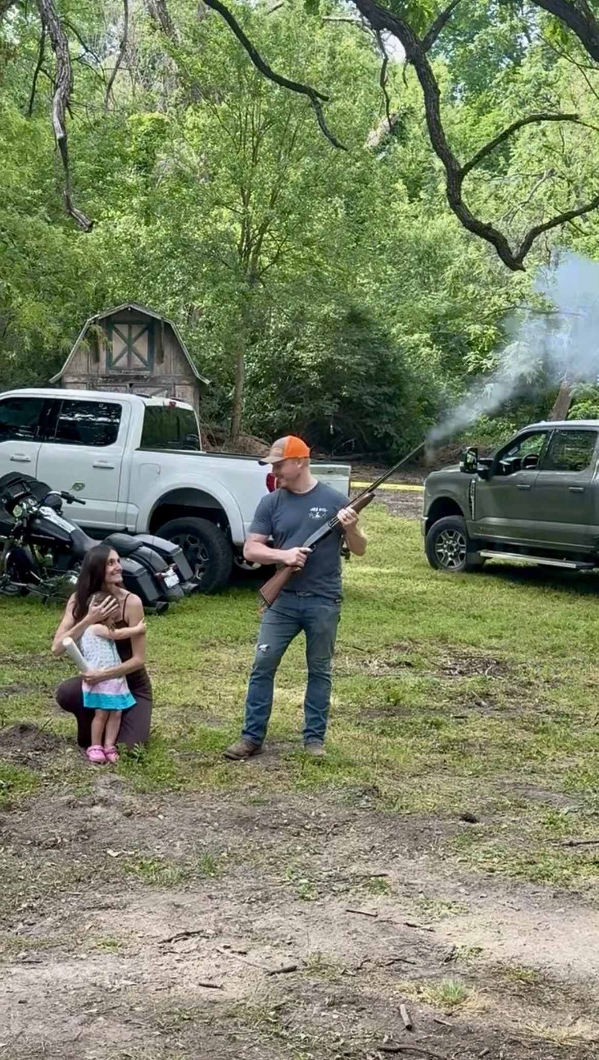 A man holding a shotgun with smoke coming from it, talking to a young girl who appears happy and is smiling, in an outdoor area with vehicles and trees in the background.