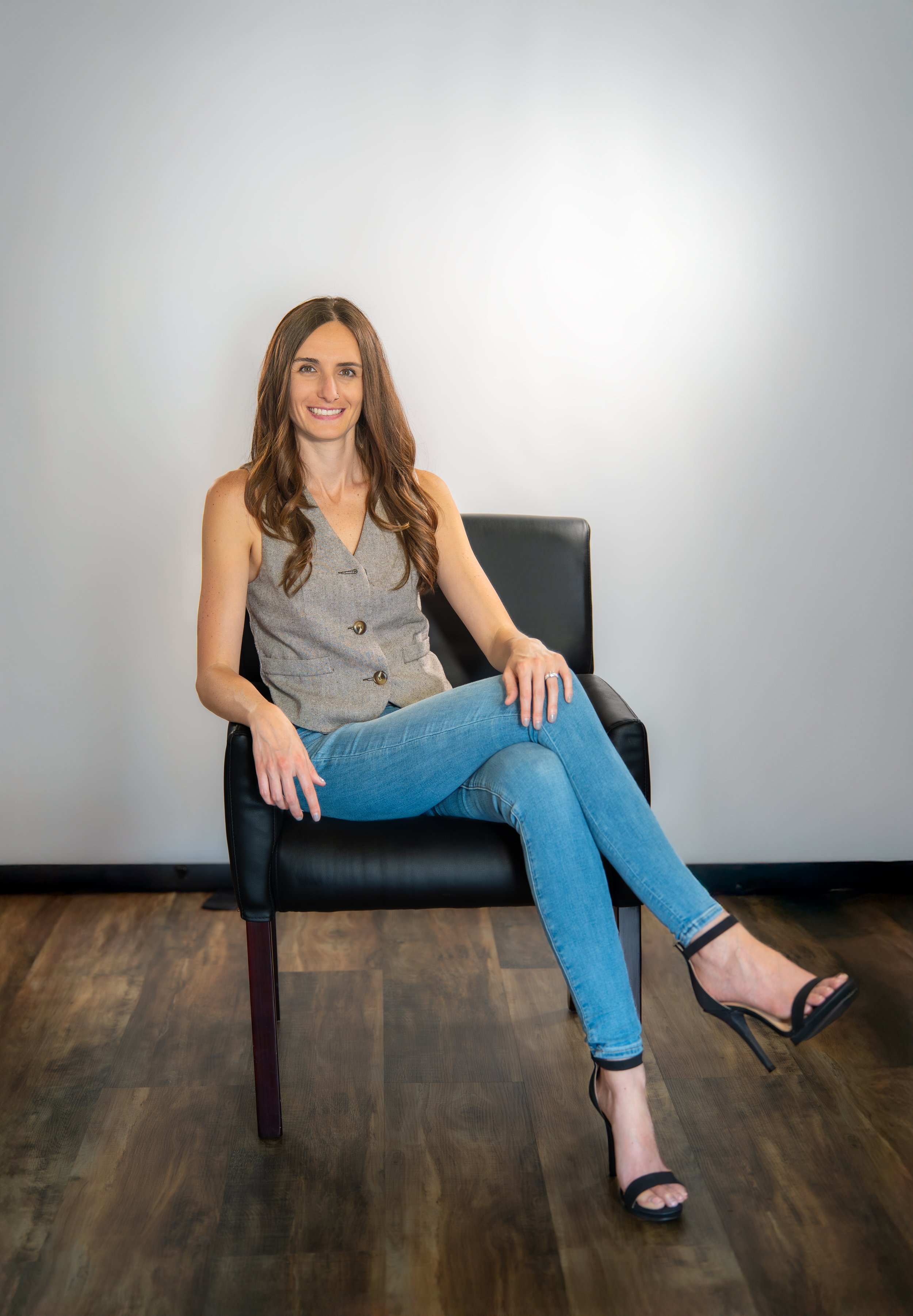 A woman with long brown hair, smiling, sitting on a black chair against a plain light gray wall. She is wearing a sleeveless gray vest, blue jeans, and black high-heeled sandals, with her legs crossed.