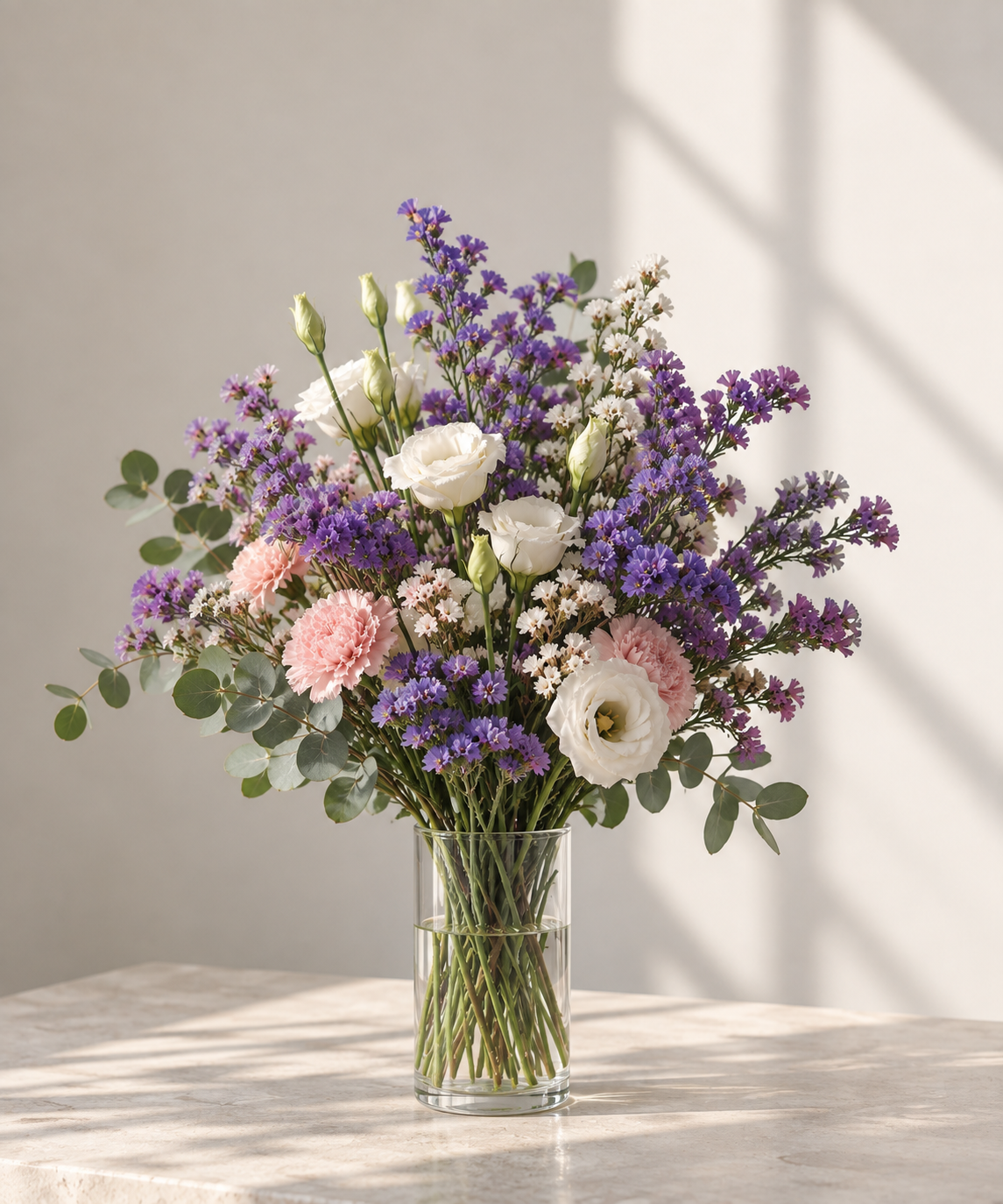 Ramo de flores en jarrón con lisianthus blancos, gladiolos rosa, estatice morado y pequeñas margaritas blancas, con hojas de eucalipto, en una superficie de mármol y luz natural.