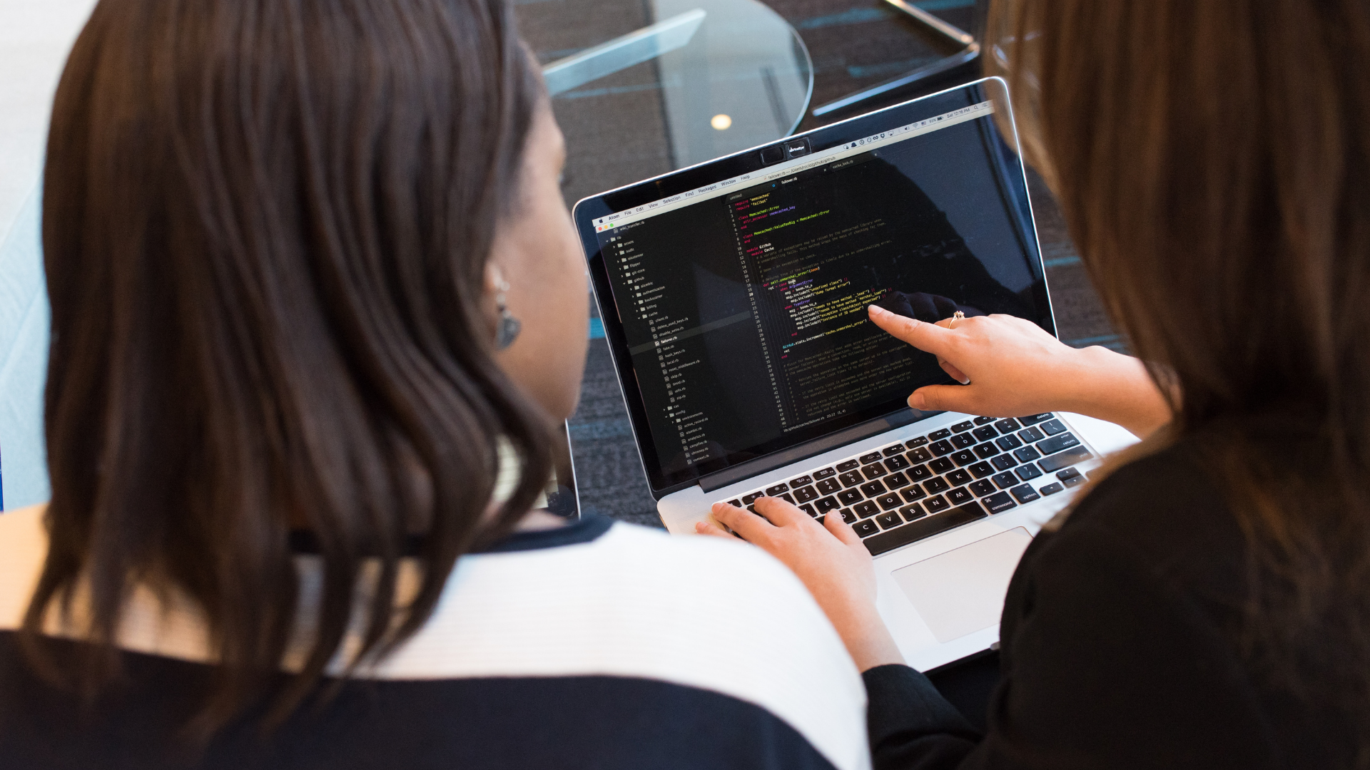 Two women sit together at a table, working on a laptop that displays lines of colorful code, suggesting a programming or coding activity.