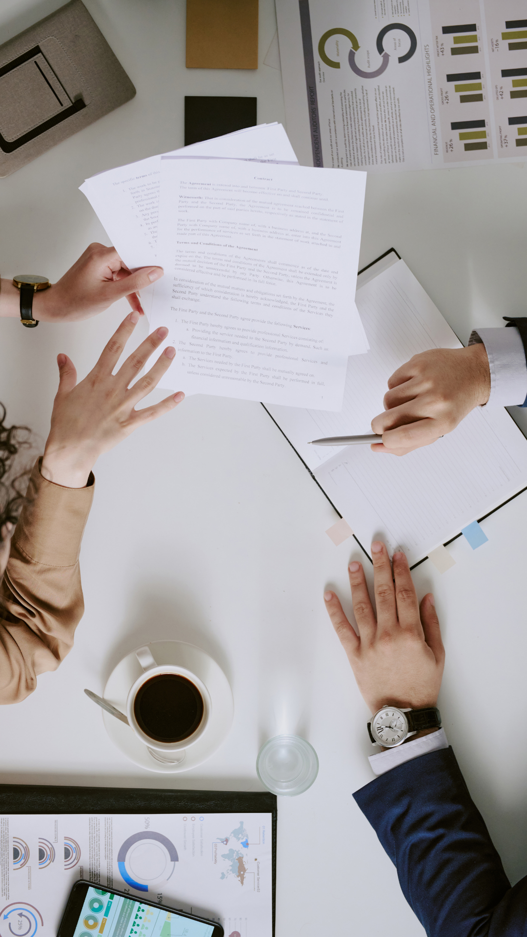 A top-down view of a business meeting with two people exchanging documents at a white table. There are charts, a smartphone, a cup of coffee, notebooks, and a gray pencil case on the table.