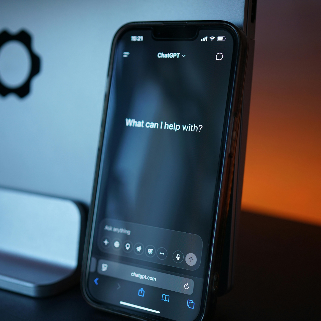 An iPhone displays the ChatGPT interface with the prompt 'What can I help with?' on the screen, placed on a stand in front of a blurred computer monitor and an orange background.