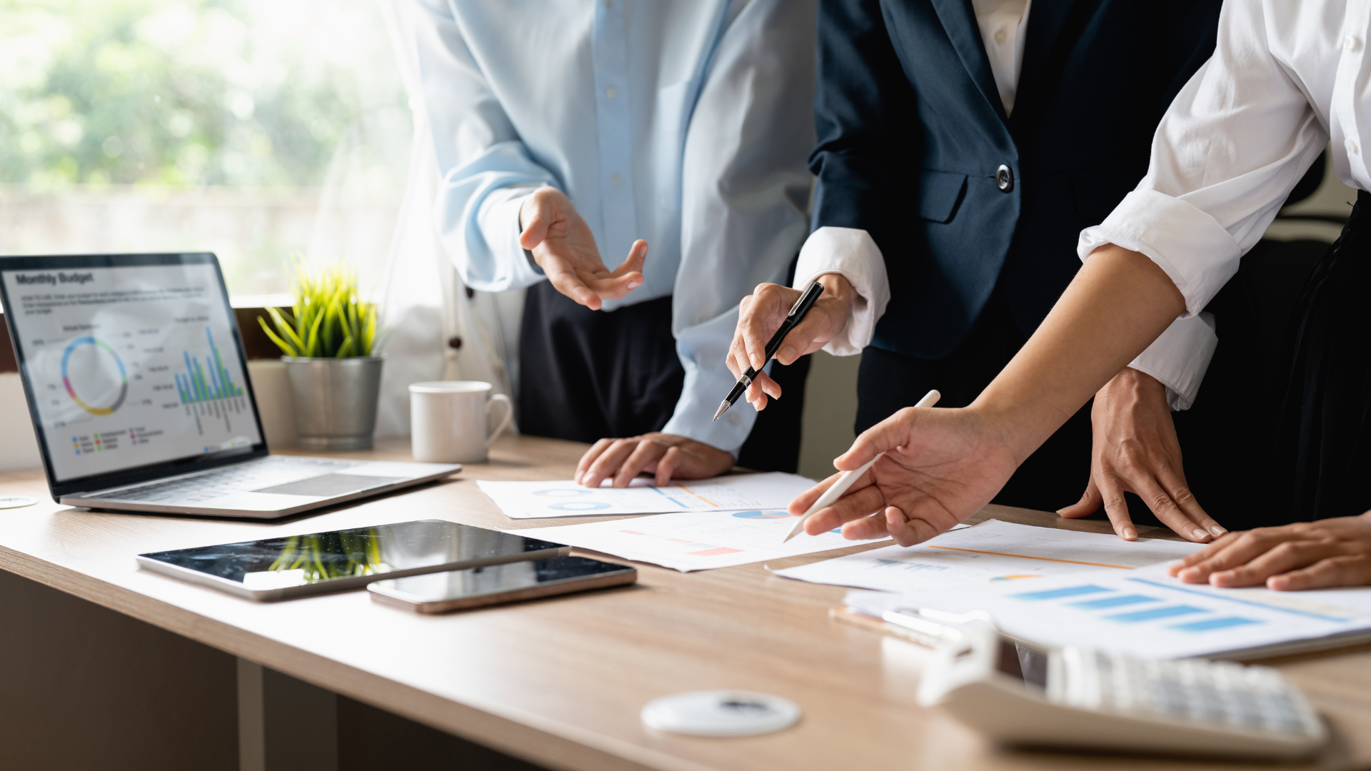 Multiple professionals in business attire collaborating and discussing documents and charts on a wooden table in a modern office with natural light.