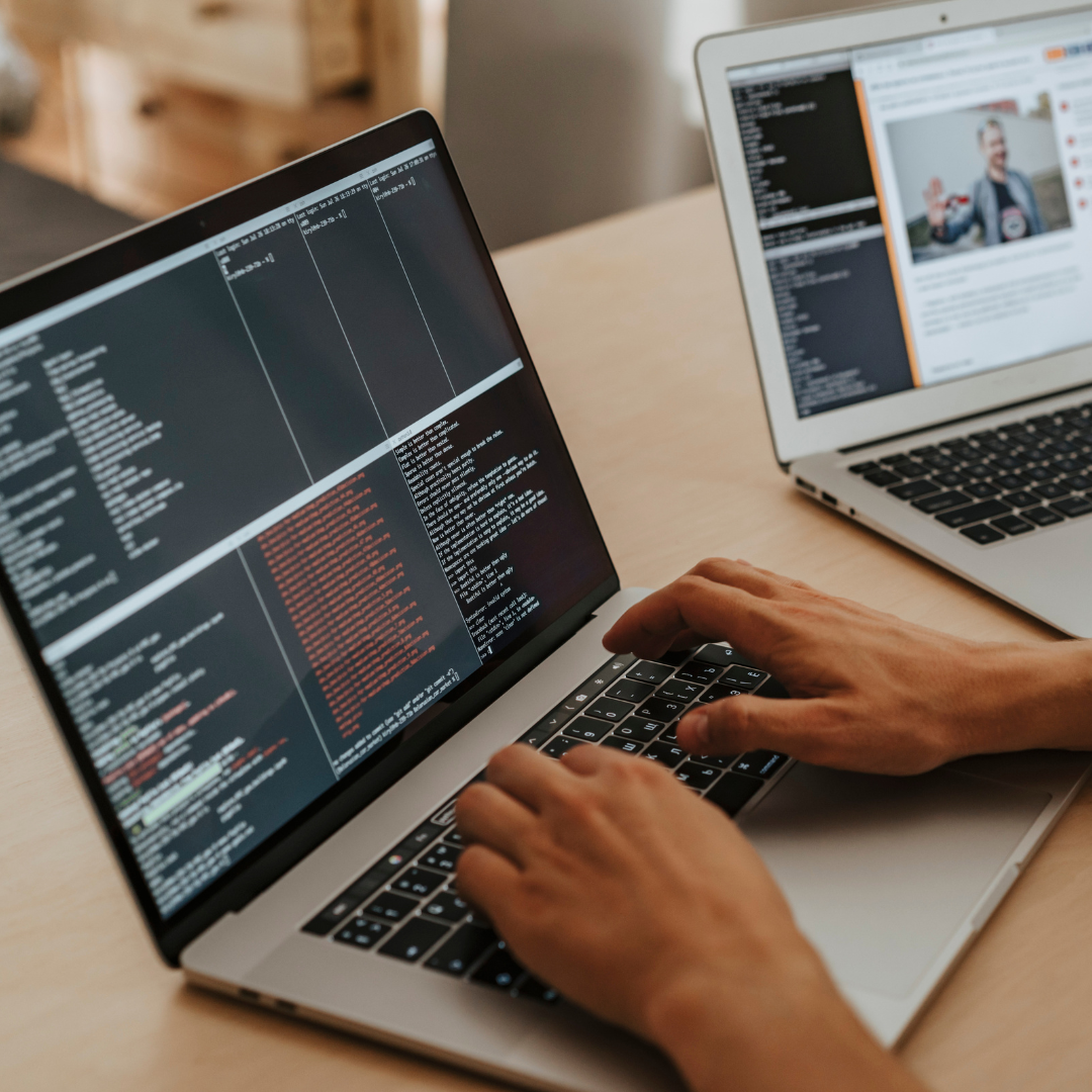 Person working on a laptop with code on the screen, next to another open laptop showing a web page with a person giving a presentation.