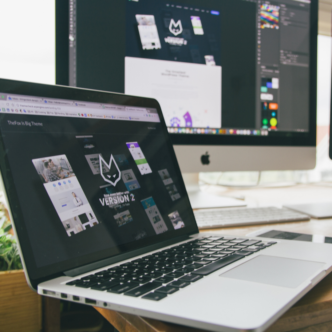 A MacBook open on a desk showing a website about a design theme, with a desktop monitor in the background also displaying design work in graphic design software.