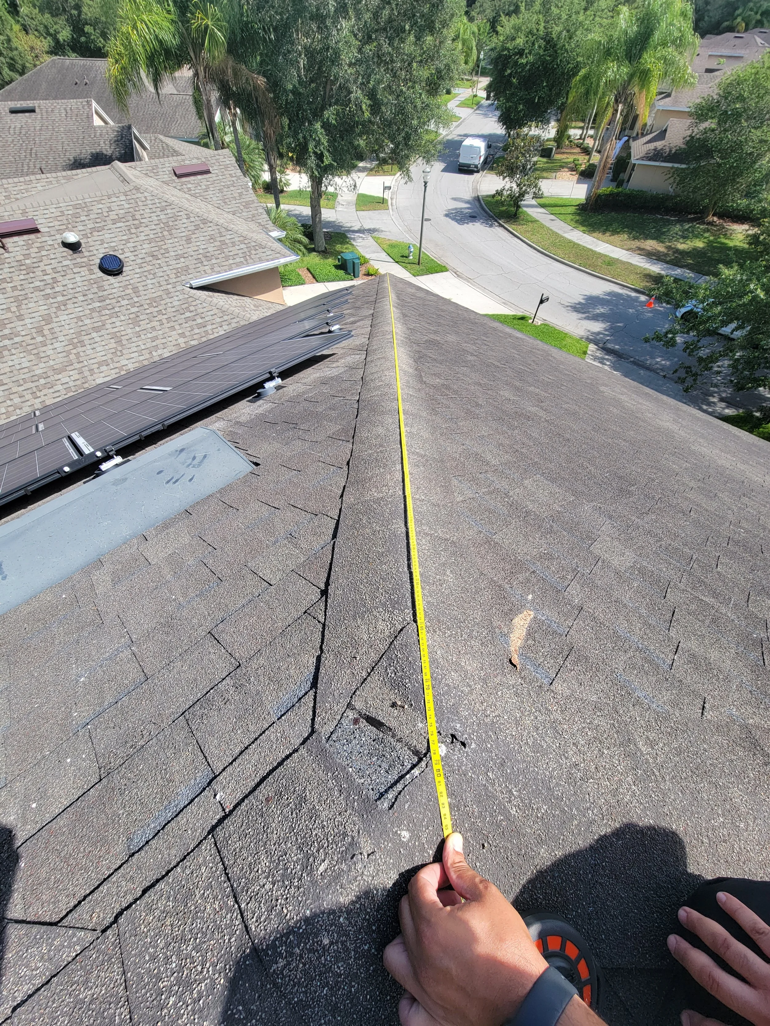 Person measuring roof shingles with a tape measure from the peak of a residential roof, showing shingles, trees, and a suburban neighborhood street below.