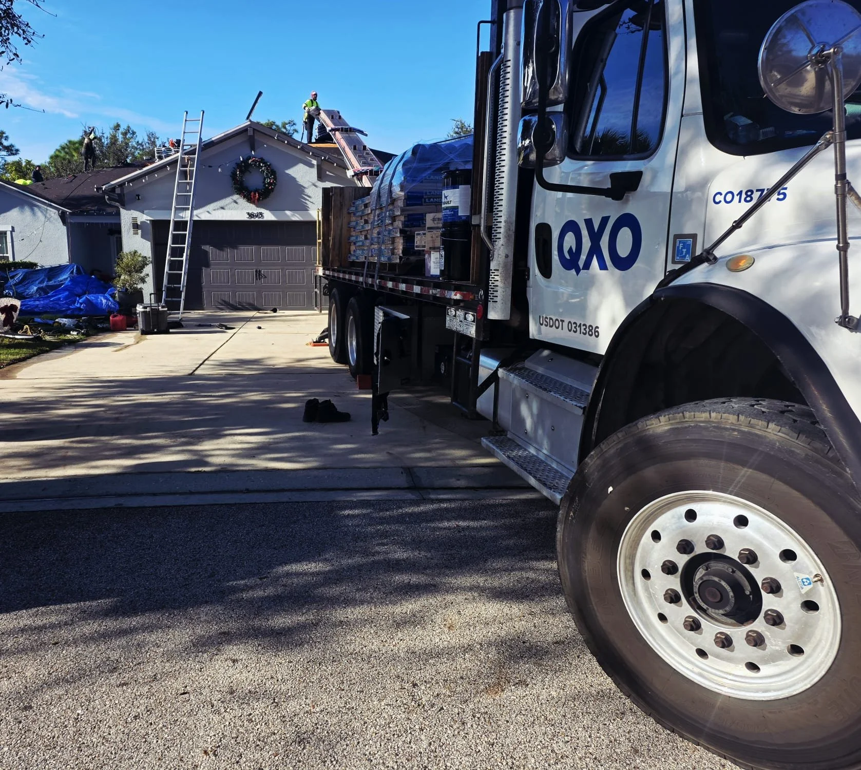 A white delivery truck parked in front of a house with house number 36473, with a large load of bricks or tiles on its flatbed, and workers on the roof installing solar panels. The house is decorated with a Christmas wreath, and a ladder leans against the roof. The driveway has tools and supplies, and a blue tarp covers some items on the ground.