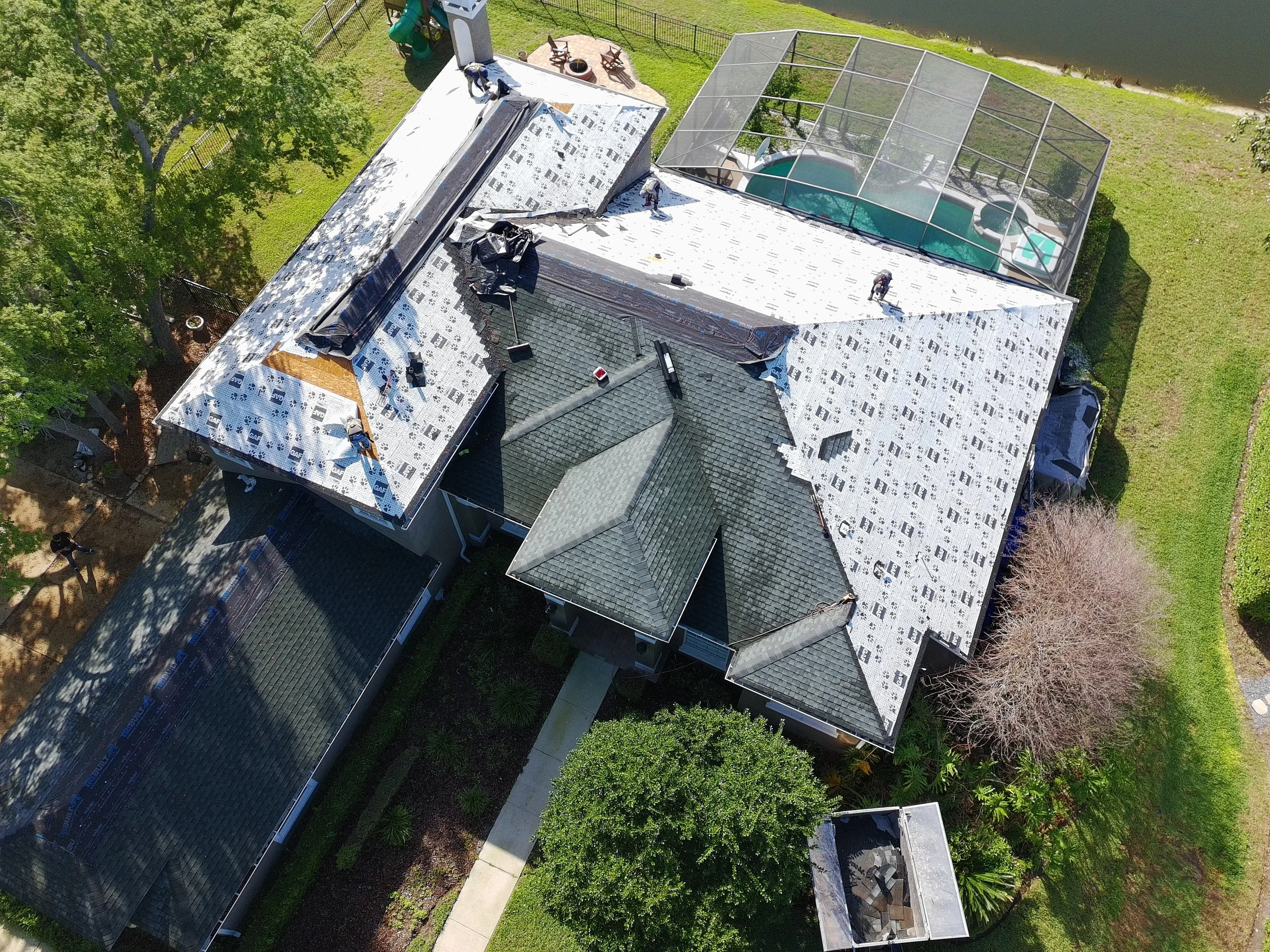 Aerial view of a house under construction with workers installing roofing material. The house has a tiled roof section, a covered outdoor pool area, and nearby water body, with trees and a fence around the yard.