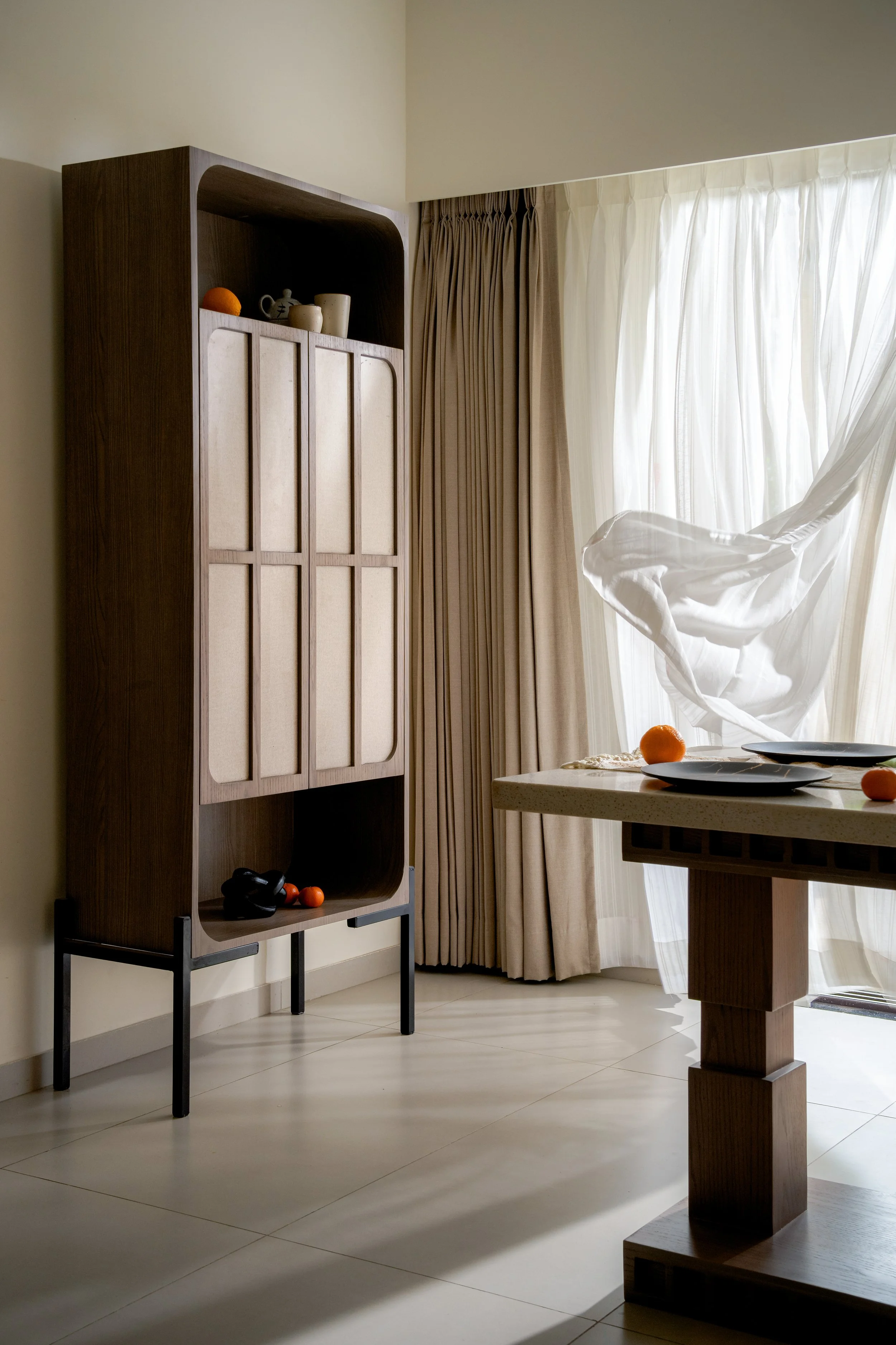 A corner of a room with a wooden cabinet on a stand, a round table with black plates and oranges, and beige curtains covering a window with sunlight.