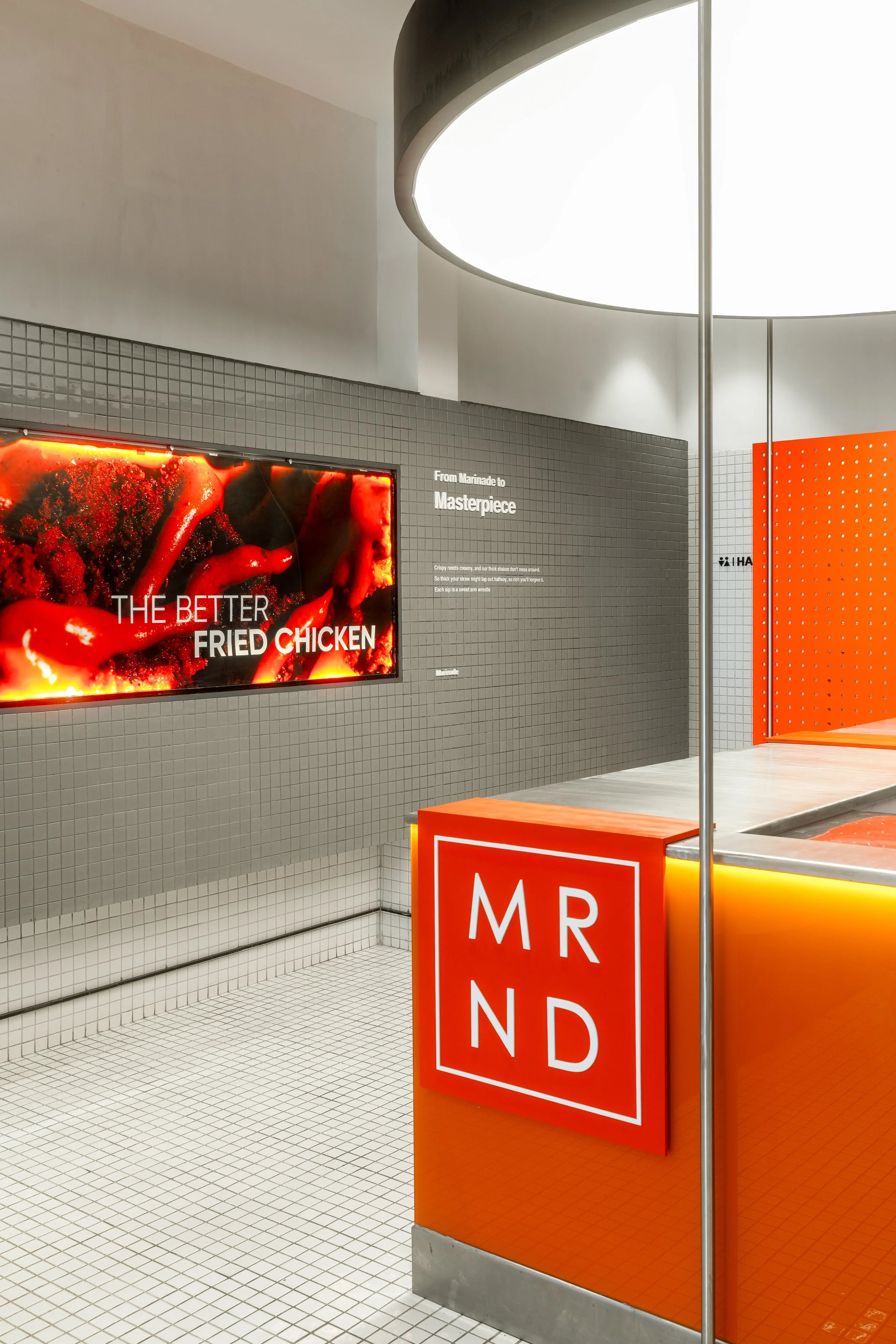 Interior view of a modern restaurant or food court with a large illuminated circular ceiling light, gray tiled wall with a digital sign advertising fried chicken, orange and gray decor with signs labeled 'Order' and 'Pickup'.