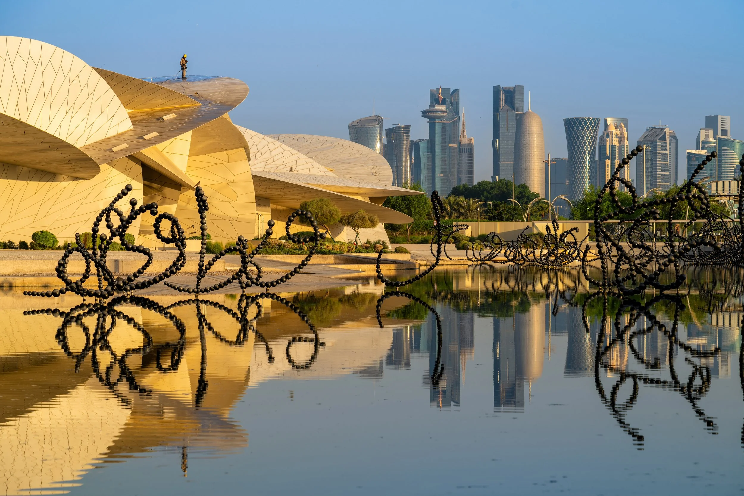 A construction worker standing on top of a modern, golden architectural structure in the foreground, with a city skyline featuring tall, contemporary skyscrapers in the background, reflected in a body of water.