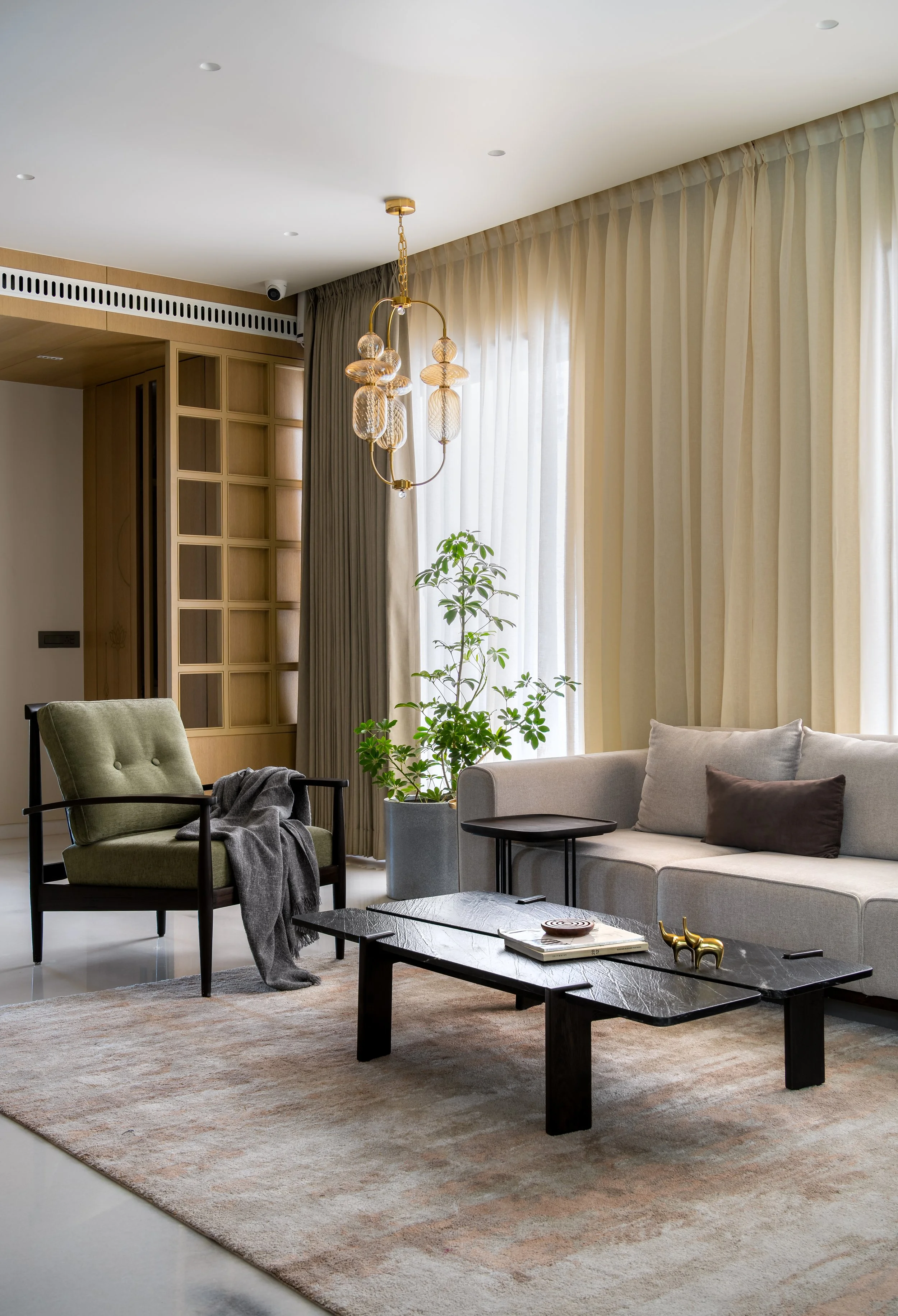 Living room with beige curtains, a light-colored sofa, a green armchair with a blanket, two black coffee tables, a potted plant, a gold and glass chandelier, and a textured beige rug.