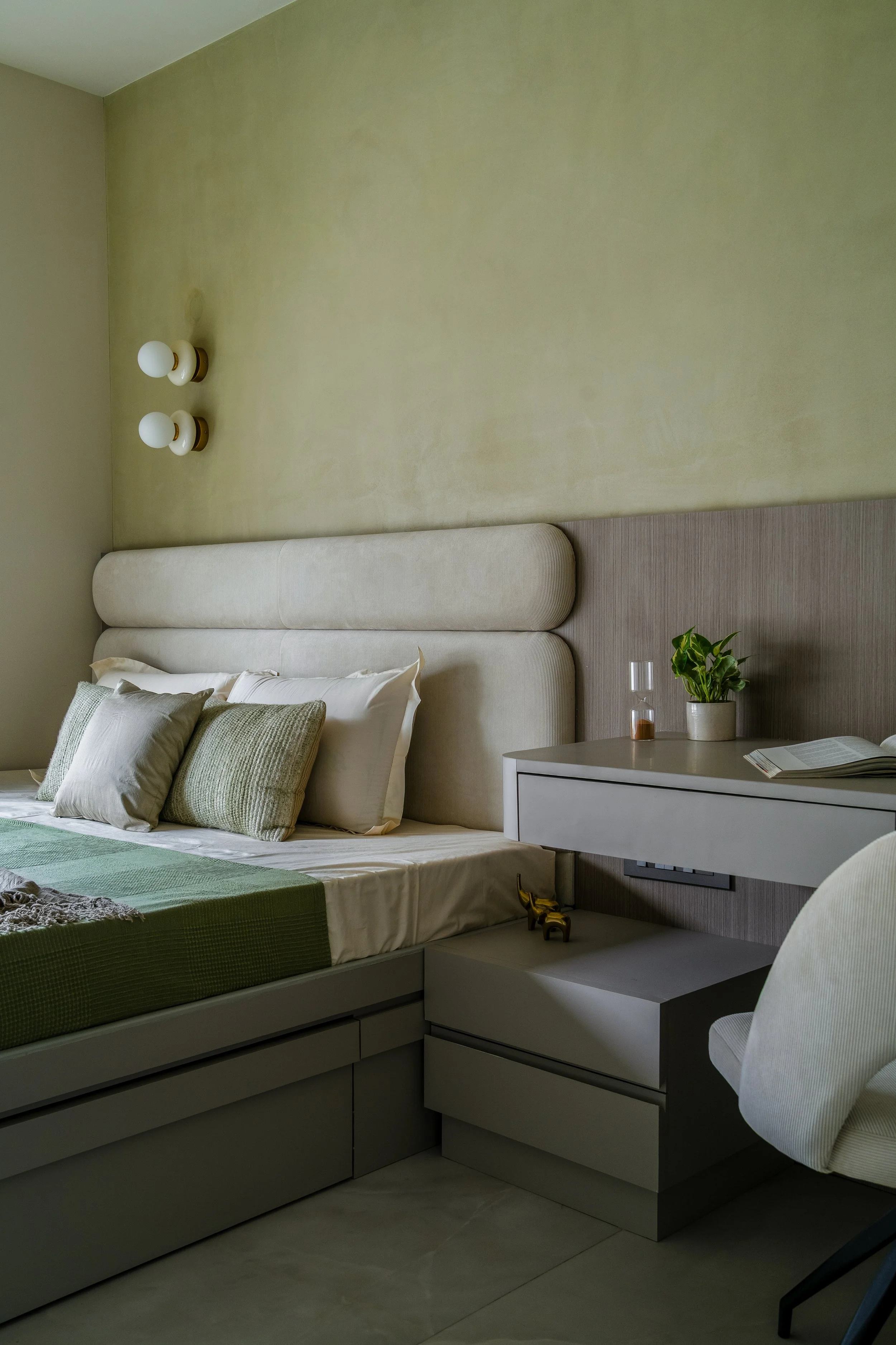 A bedroom with a bed, pillows, a nightstand, a small lamp, a potted plant, a glass, and a book, with a beige headboard and light-colored walls.