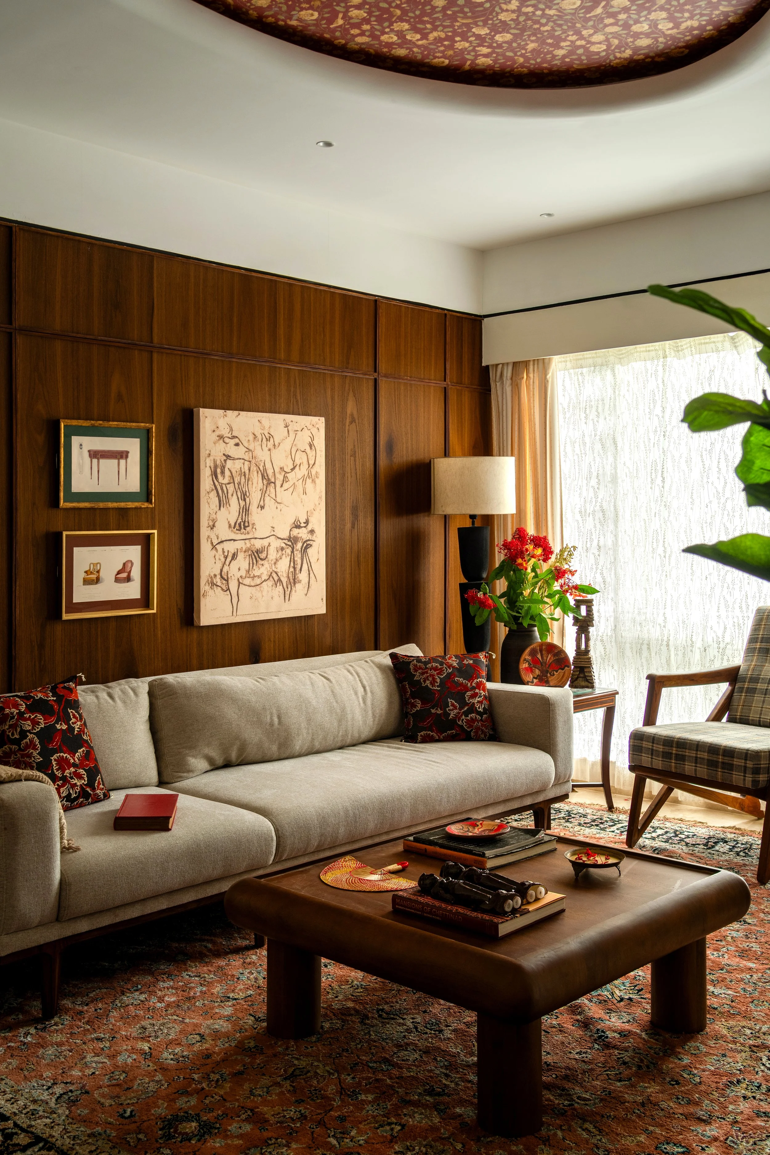 Living room with a beige sofa, black and red floral throw pillows, wooden coffee table with books and decorative objects, wood-paneled wall with framed artwork, table lamp, and a large window with sheer curtains. Accents include a potted plant and an
