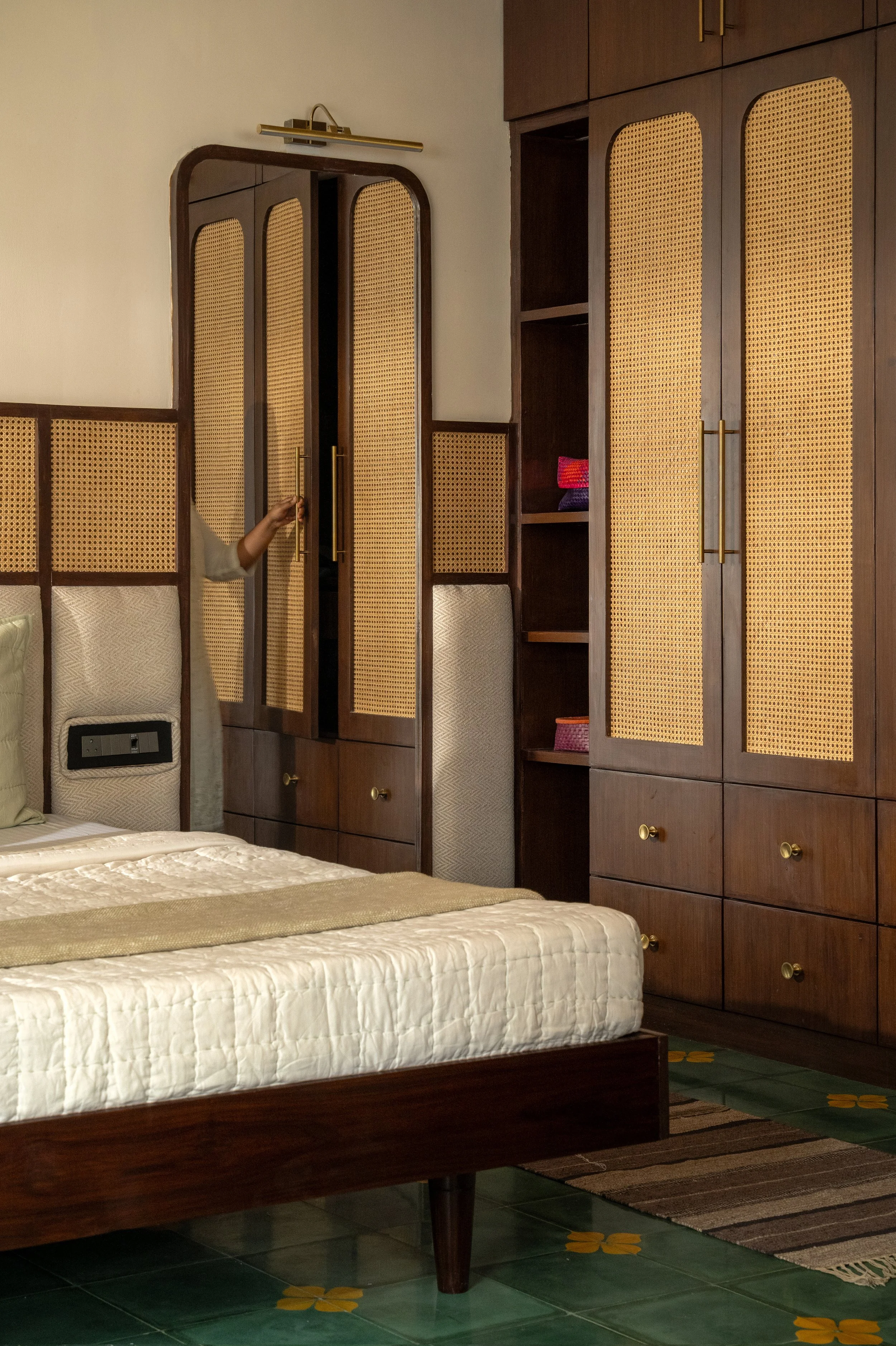 A bedroom with a bed and built-in wooden cabinets with rattan panel doors. The bed has a white quilt and a pillow, and the furniture is a dark wood. There is a decorative rug on the green-tiled floor with yellow floral patterns.