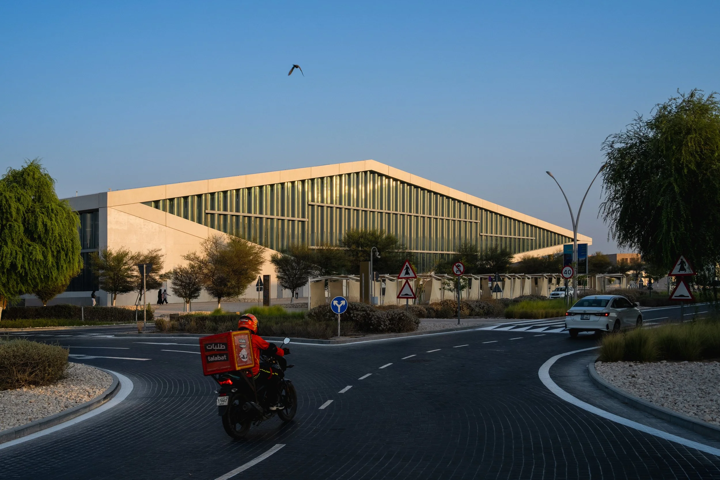 A modern building with a sloped roof made of vertical metal slats, surrounded by trees and street signs. A motorcycle rider in a red uniform is riding on a curved road near a parked silver car and traffic signs.