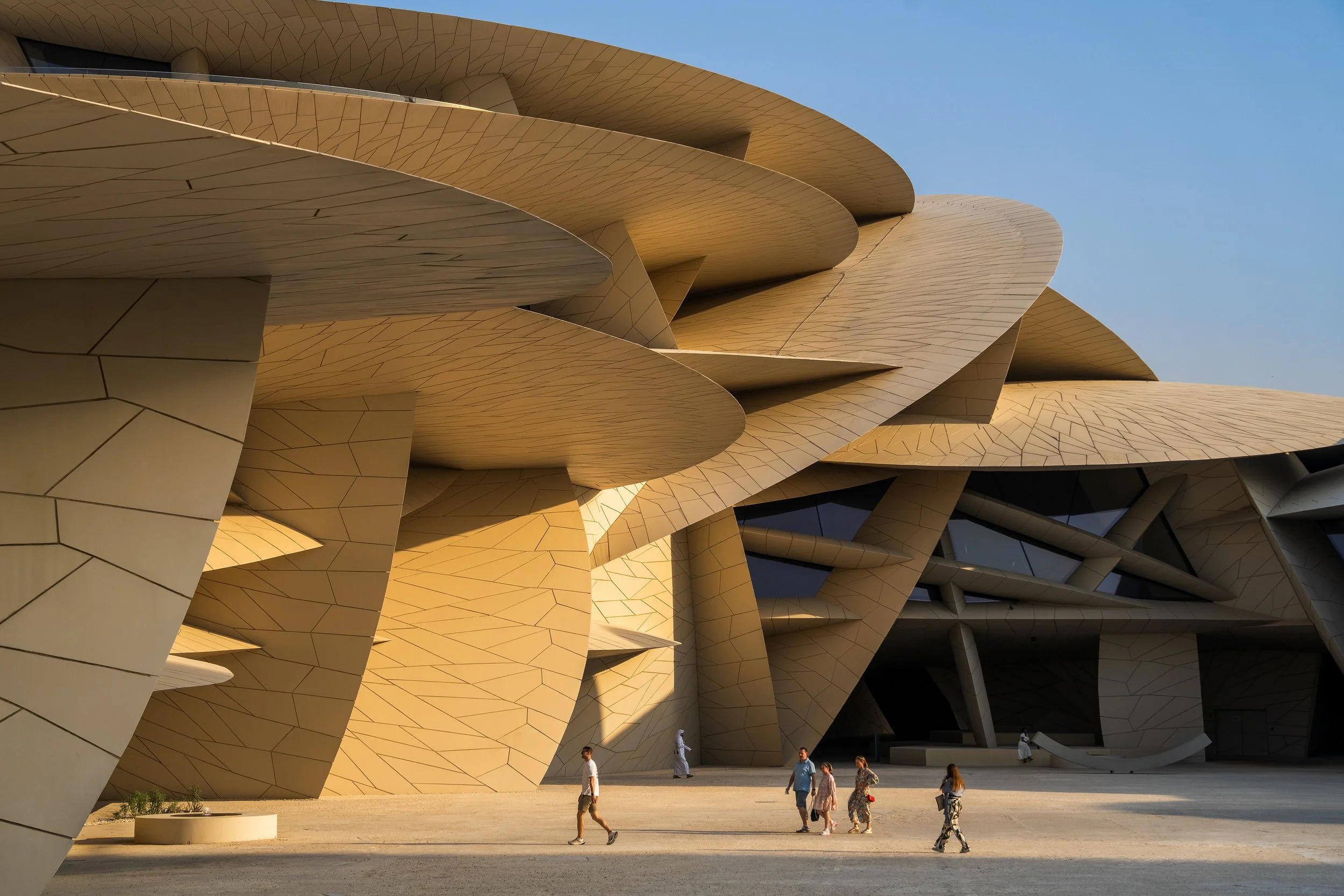 People walking near a modern architectural building with large, curved, beige panels resembling overlapping petals or sails set against a clear blue sky.