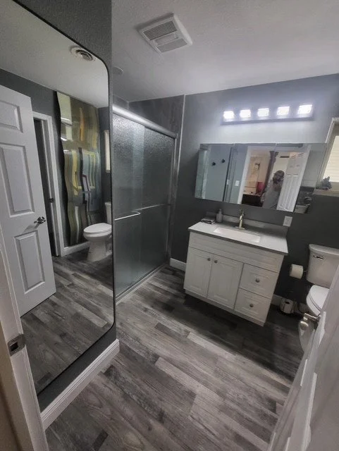 Bathroom with a glass shower, white vanity with sink, mirror, toilet, and decorative wall art.