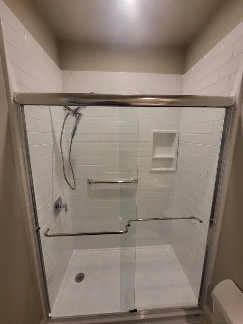 Inside a walk-in shower with white tile walls, a silver shower head on a flexible hose, a glass door, and built-in corner shelves