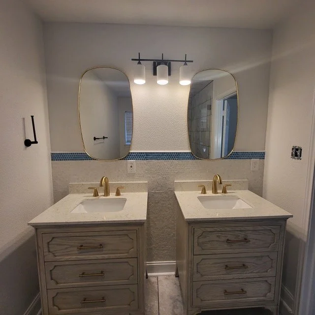 Bathroom vanity area with two sinks, gold faucets, two oval mirrors with gold trim, and a three-light modern fixture above.