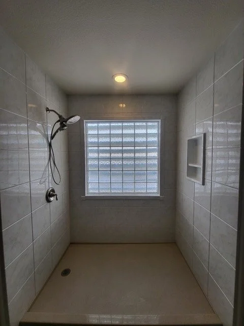 A walk-in shower with beige tile walls, a handheld showerhead, a small recessed shelf on the right wall, a frosted glass block window, and a ceiling light.