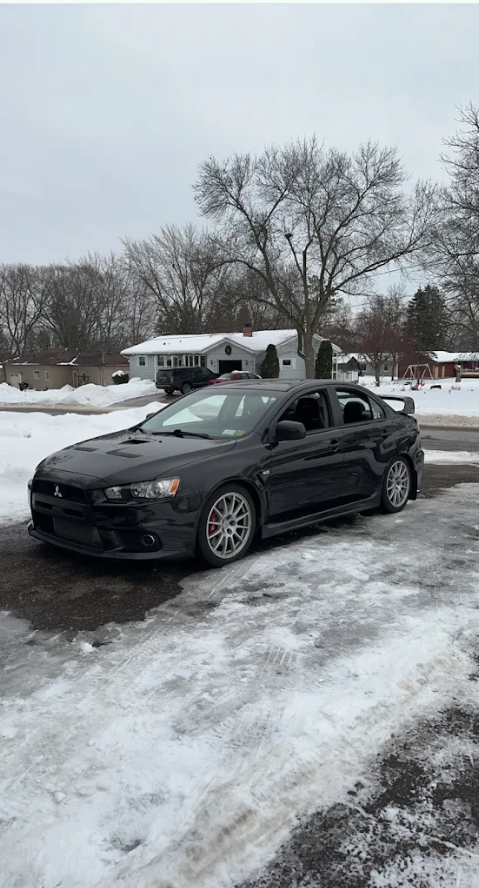Black Mitsubishi Lancer parked on snowy driveway with houses and leafless trees in the background.
