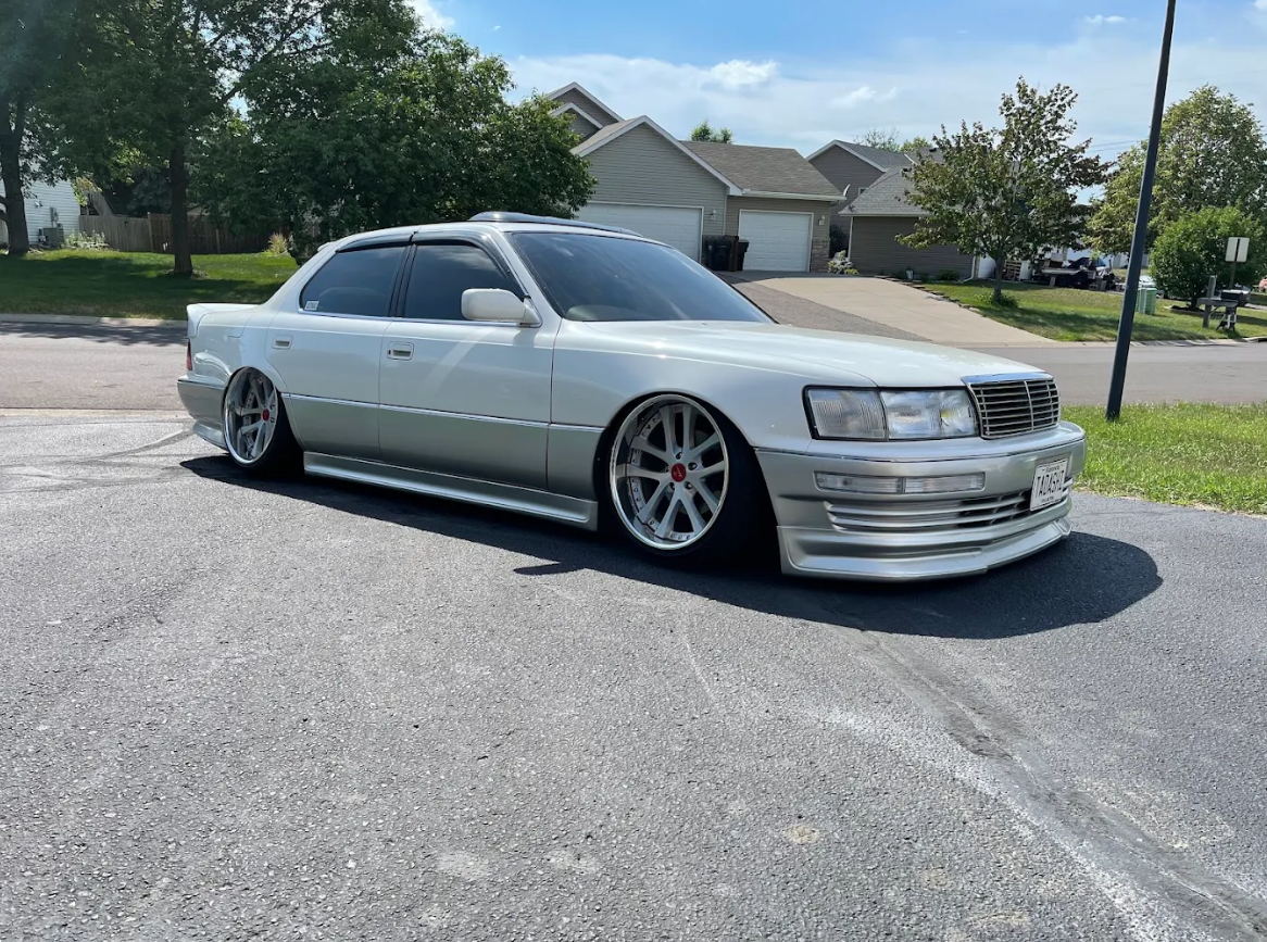 Silver modified sedan parked on the street in a residential neighborhood with single-family homes, trees, and a mailbox visible in the background.