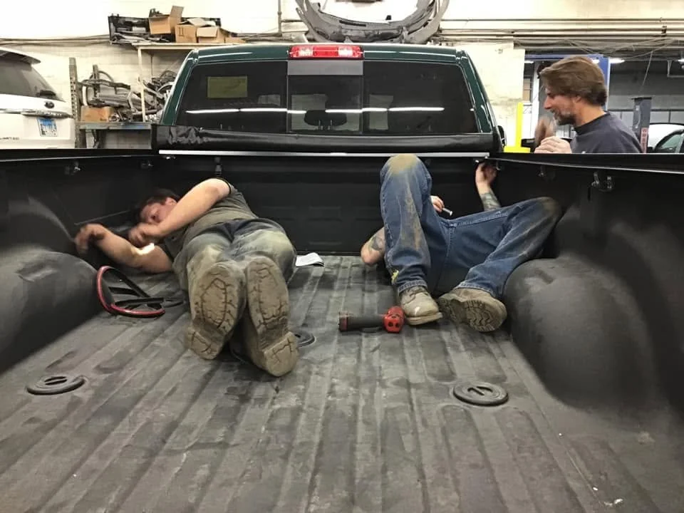Two men are lying inside the bed of a pickup truck, working on repairs, with a man standing outside the truck on the right, holding a tool and talking to them in a workshop.