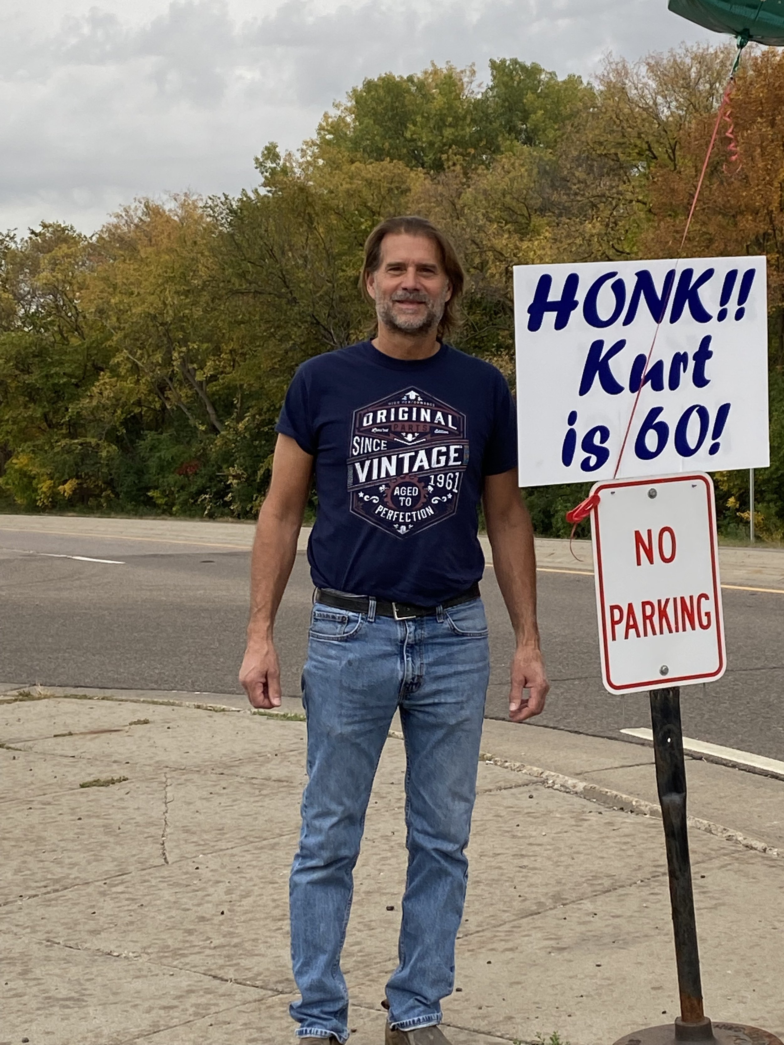 A middle-aged man with brown hair and a beard standing outdoors next to a sign that reads 'HONK!! Kart is 60!' on a cloudy day. He is wearing a navy T-shirt with a vintage graphic and blue jeans.
