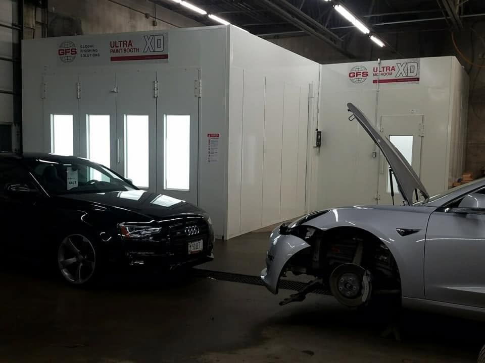 A black Audi and a silver Tesla parked inside an automotive repair shop, with the Tesla's front end disassembled and an open hood.