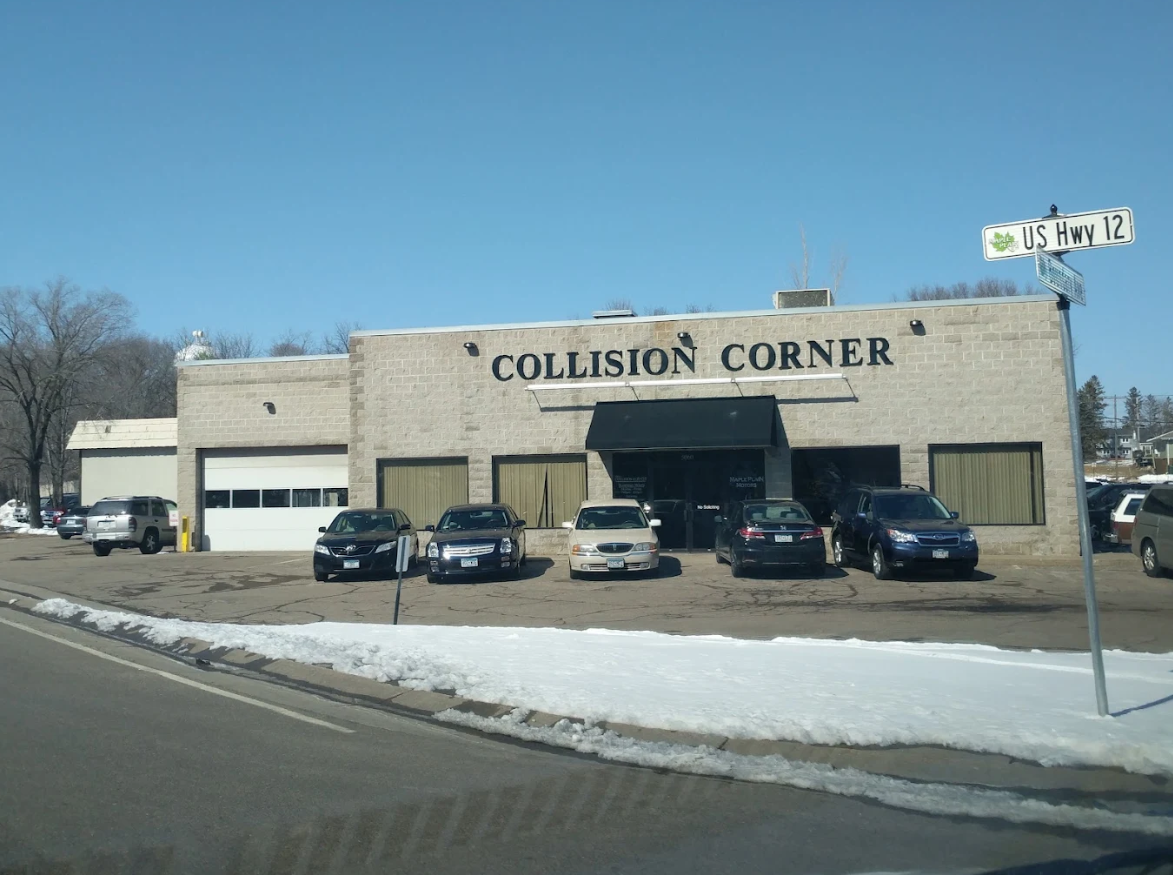 A car repair shop named 'Collision Corner' with a beige brick exterior, parking lot with several cars, and a sign indicating U.S. Highway 12. Snow is present on the ground.