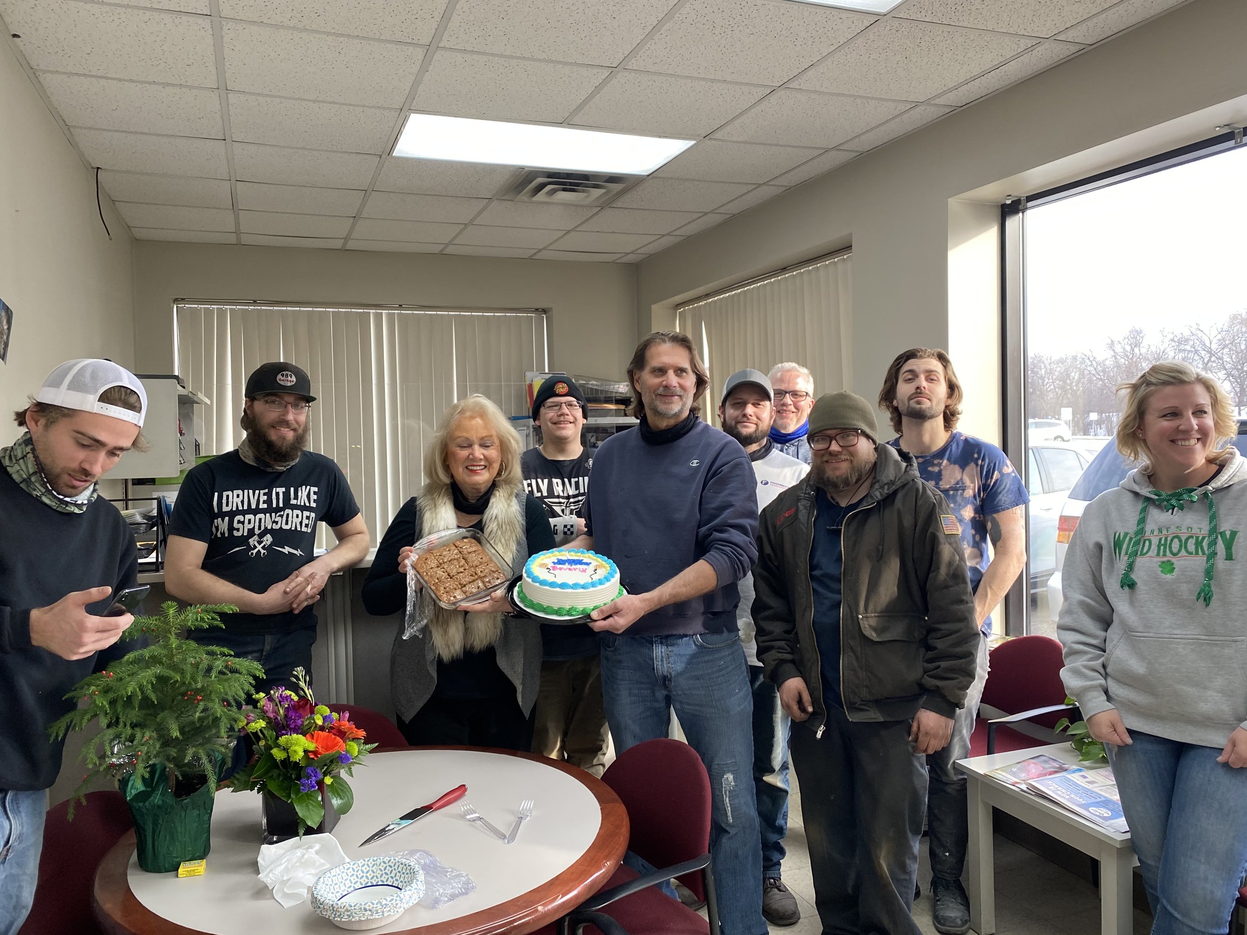 A group of people gather indoors for a celebration, with a man holding a decorated cake and a woman holding a dish. There are flowers on the table and a window with blinds in the background.