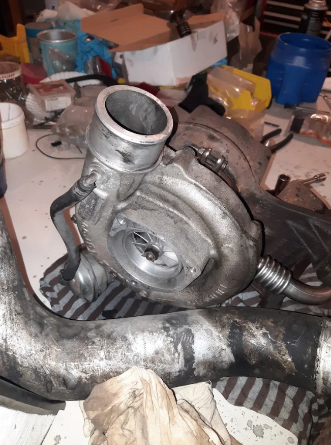 A turbocharger with pipes and components on a cluttered workbench in a garage or workshop.